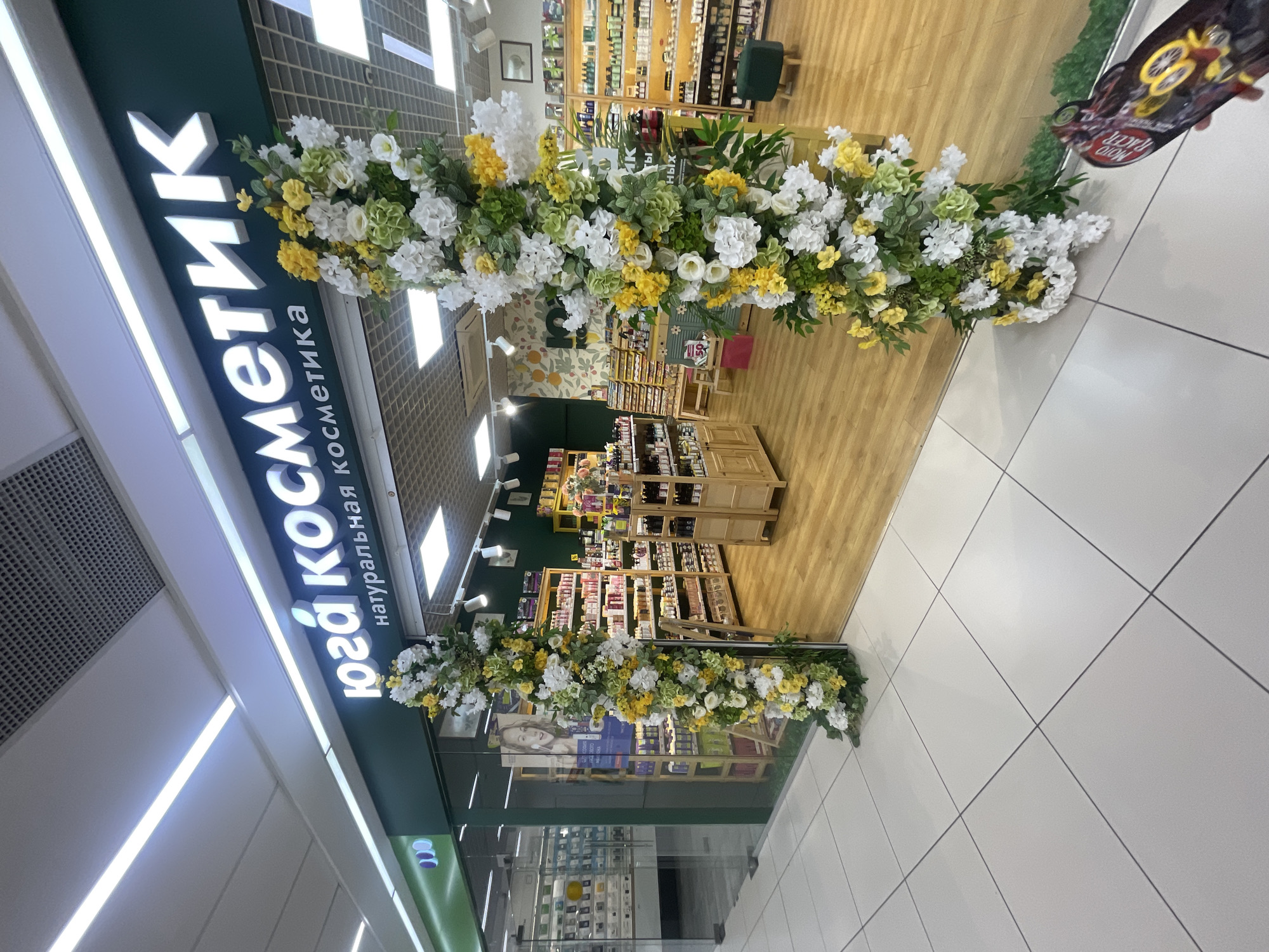 The image shows the entrance to a "юга Косметика" store, which translates to "South Cosmetics," specializing in natural cosmetics. The store is located in Sochi, Russia, based on the Cyrillic text. The storefront is decorated with a floral archway of white, yellow, and green flowers and greenery. The interior is visible, with shelves stocked with products. The floor is tiled, and the perspective suggests the photo was taken inside a shopping mall or similar indoor space.