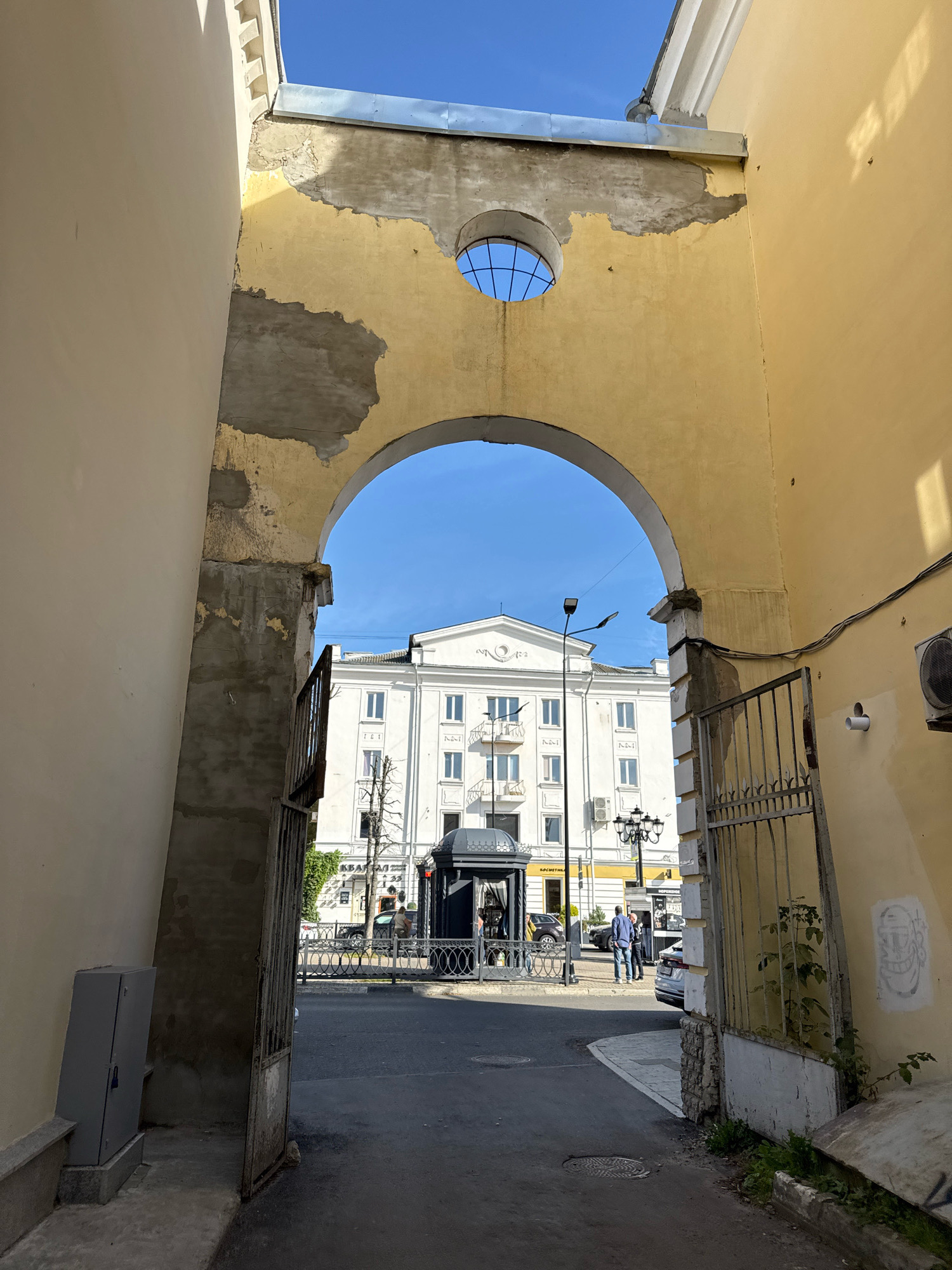 An archway frames a view of a white building and a street in Tver, Russia. The archway itself is made of weathered yellow stucco, with patches of concrete showing through and a circular window with a grid at the top. A metal gate stands open to the right of the archway. On the street, two men in casual clothing are visible, and several cars are parked. A black kiosk with a silver railing is also present on the sidewalk. The sky is clear and blue. The overall impression is one of a somewhat aged urban environment.