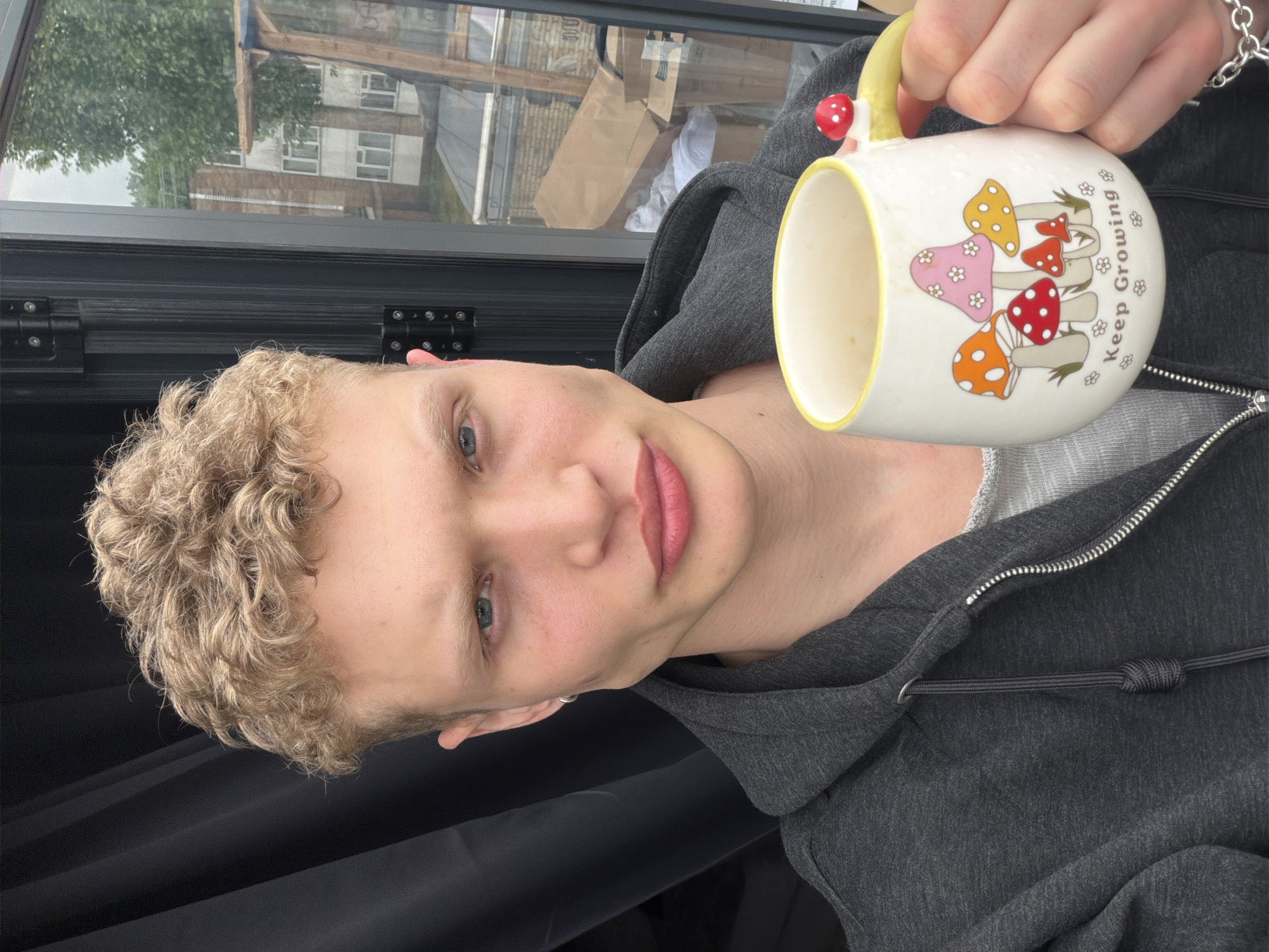 A young person with short, curly blonde hair, wearing a grey hooded sweatshirt, is holding a ceramic mug in their right hand. The mug is white with a yellow interior rim and handle. There are several colorful mushroom designs on the mug and the phrase "Keep Growing" written next to them. The handle has a small red mushroom attached. The person is looking directly at the camera with a slight smile. The background appears to be an interior space with a window showing an exterior view of buildings and trees. Based on the architecture visible through the window and general aesthetic, the location is potentially somewhere in the city of London, United Kingdom.