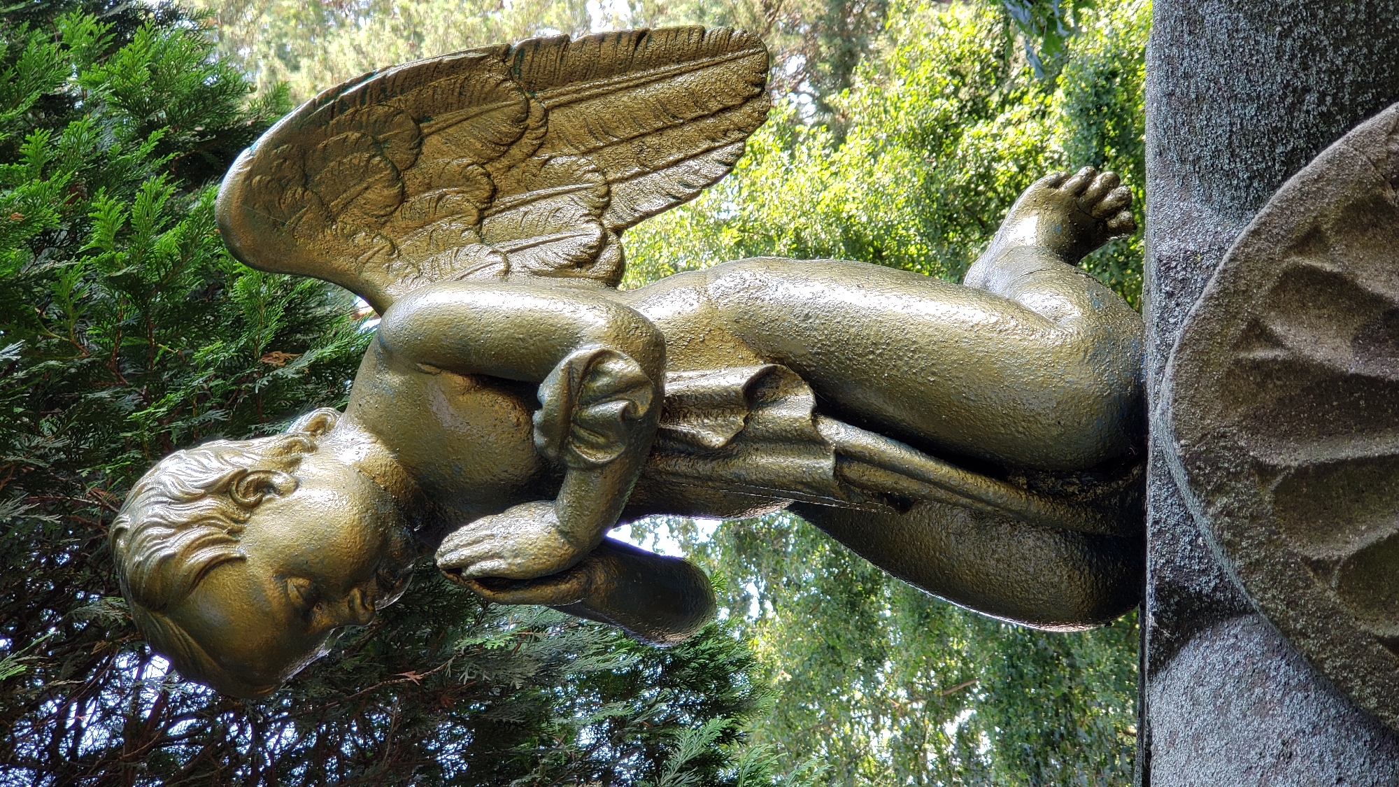 This is a full, low-angle outdoor vertical shot of a weathered, gold-toned statue of a child with angel wings, kneeling with hands clasped in prayer. The statue is positioned on top of a grey, intricately carved stone structure, possibly a monument or tomb. Lush green foliage, likely trees, forms the background and frames the upper left portion of the statue. The statue's surface exhibits a textured, somewhat worn appearance, with hints of verdigris in some recessed areas. The lighting suggests it is daytime, with sunlight illuminating the statue from the right, casting soft shadows. This statue is located at the Kalvária cemetery in Prešov, Slovakia.