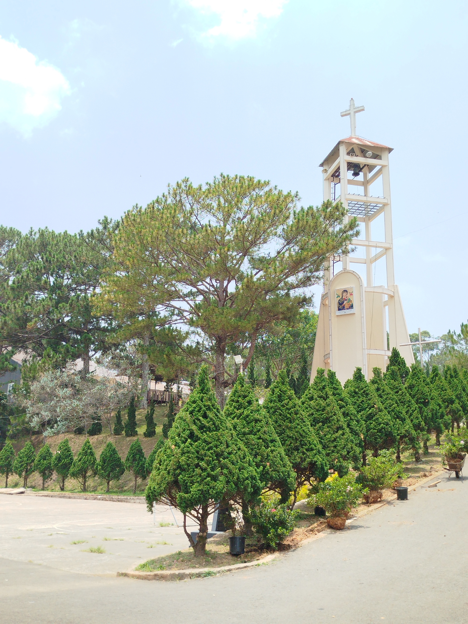 The image depicts a church bell tower in Lang Biang - Đà Lạt, Vietnam. The tower is a light-colored, multi-tiered structure topped with a Christian cross. A religious image is affixed to the tower's side.
The foreground features a variety of trees and landscaping, including rows of conical trees and various flowering plants in pots. A paved area or road stretches across the bottom of the frame, indicating a public space. The sky is light blue with scattered clouds. The environment suggests a well-maintained and serene setting.