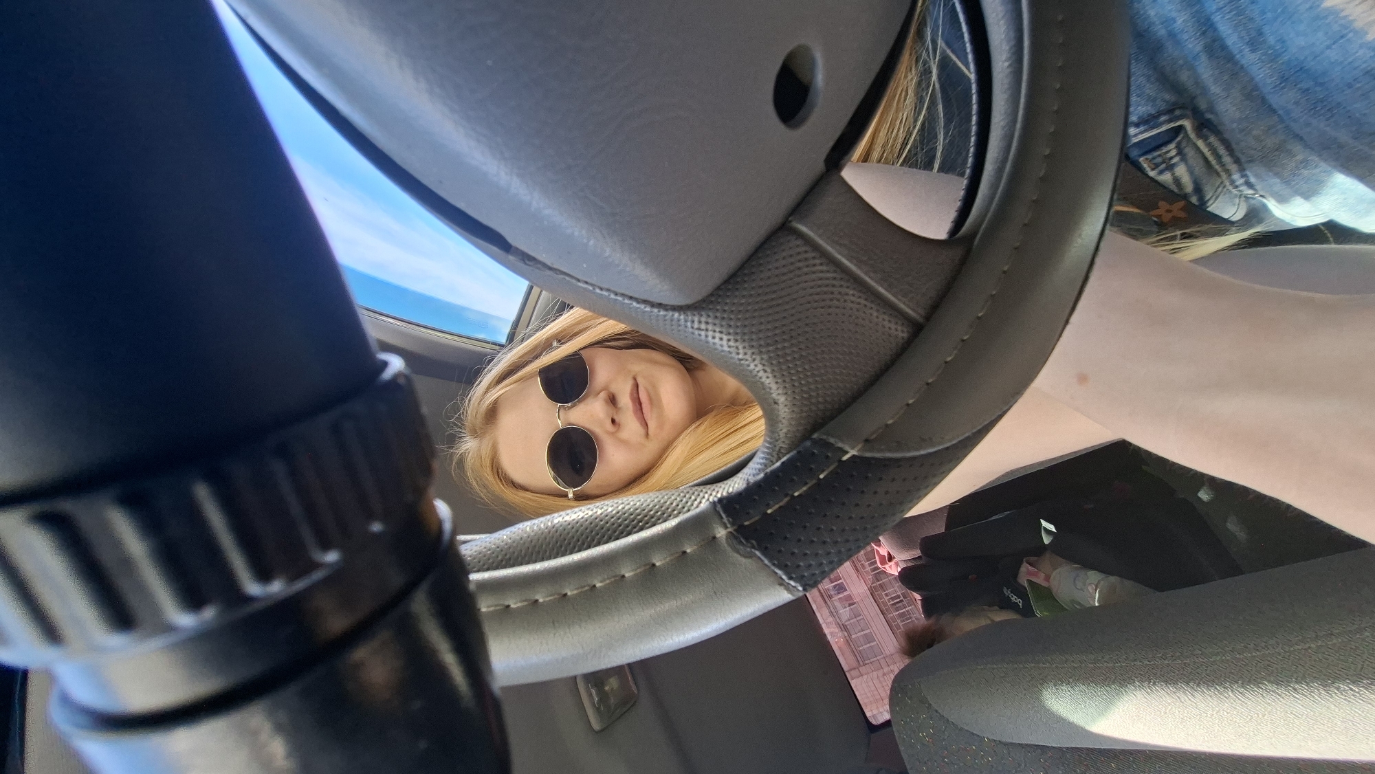 A young woman with blonde hair and sunglasses is reflected in the steering wheel of a car, looking up at the camera. She is wearing a denim jacket. The interior of the car is visible, including the steering wheel, dashboard, and seats. Sunlight streams in from outside, indicating it is daytime. The scene appears to be taking place in Chaikovsky, Russia, suggested by the provided location context. The woman's expression is neutral. No visible text is present in the image.