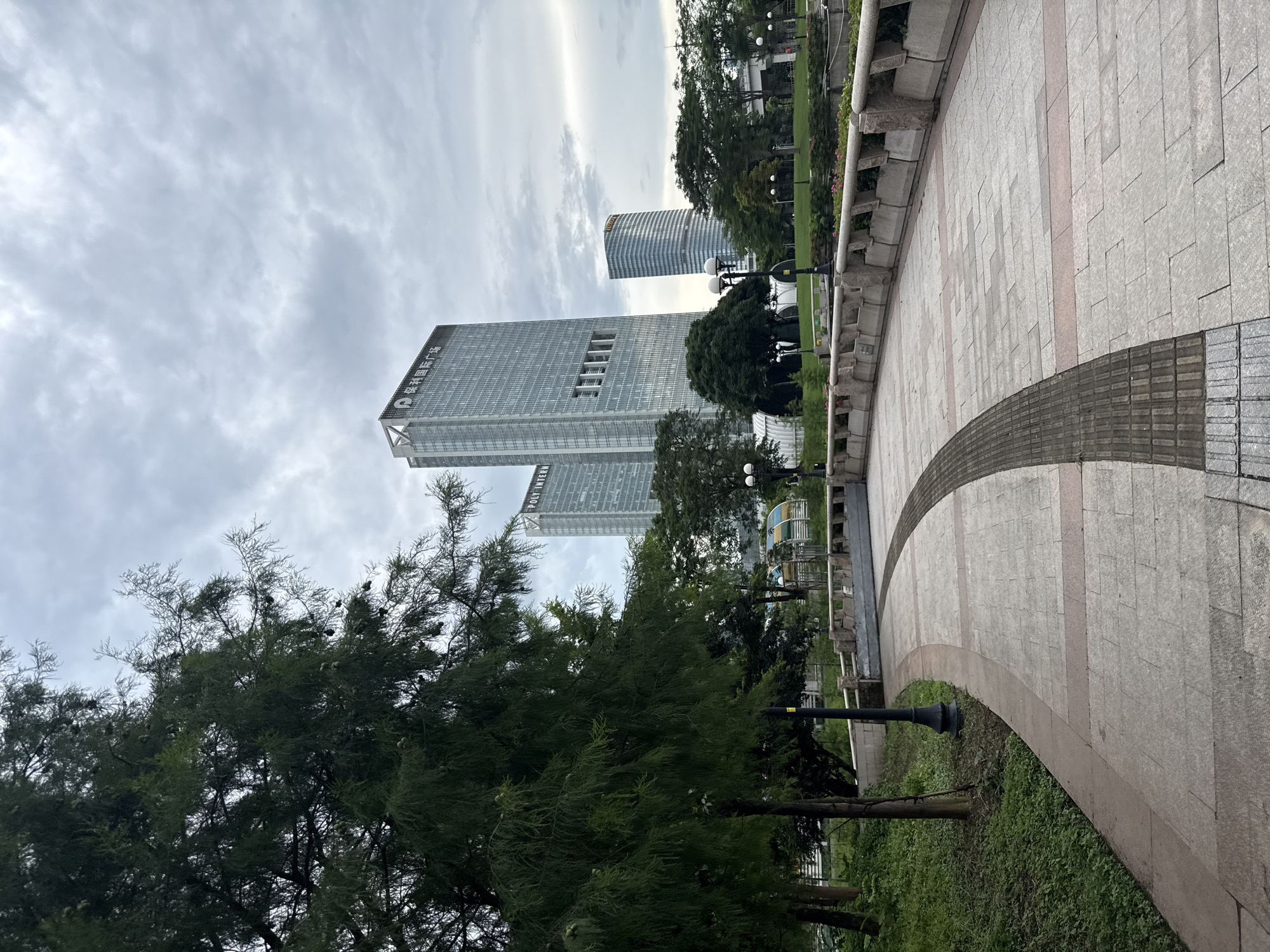 The image depicts an outdoor urban scene with modern architecture and a paved pathway. In the background, several tall glass skyscrapers dominate the skyline under a cloudy sky. One building, prominently featured, has the text "POLY INTERNATIONAL" visible on its upper section. To the left, a large, mature tree with dense foliage partially obscures the view of the buildings. In the foreground, a curved, paved pathway made of stone slabs leads into the scene. The path has a tactile paving section for visually impaired pedestrians. A low, stone wall with concrete barriers runs alongside the path. Beyond the wall, a manicured green space with trees and flower beds is visible, with lampposts scattered throughout. The location is identified as Tianhe District, China, suggesting a commercial or business district. There are no people or animals visible in the image.