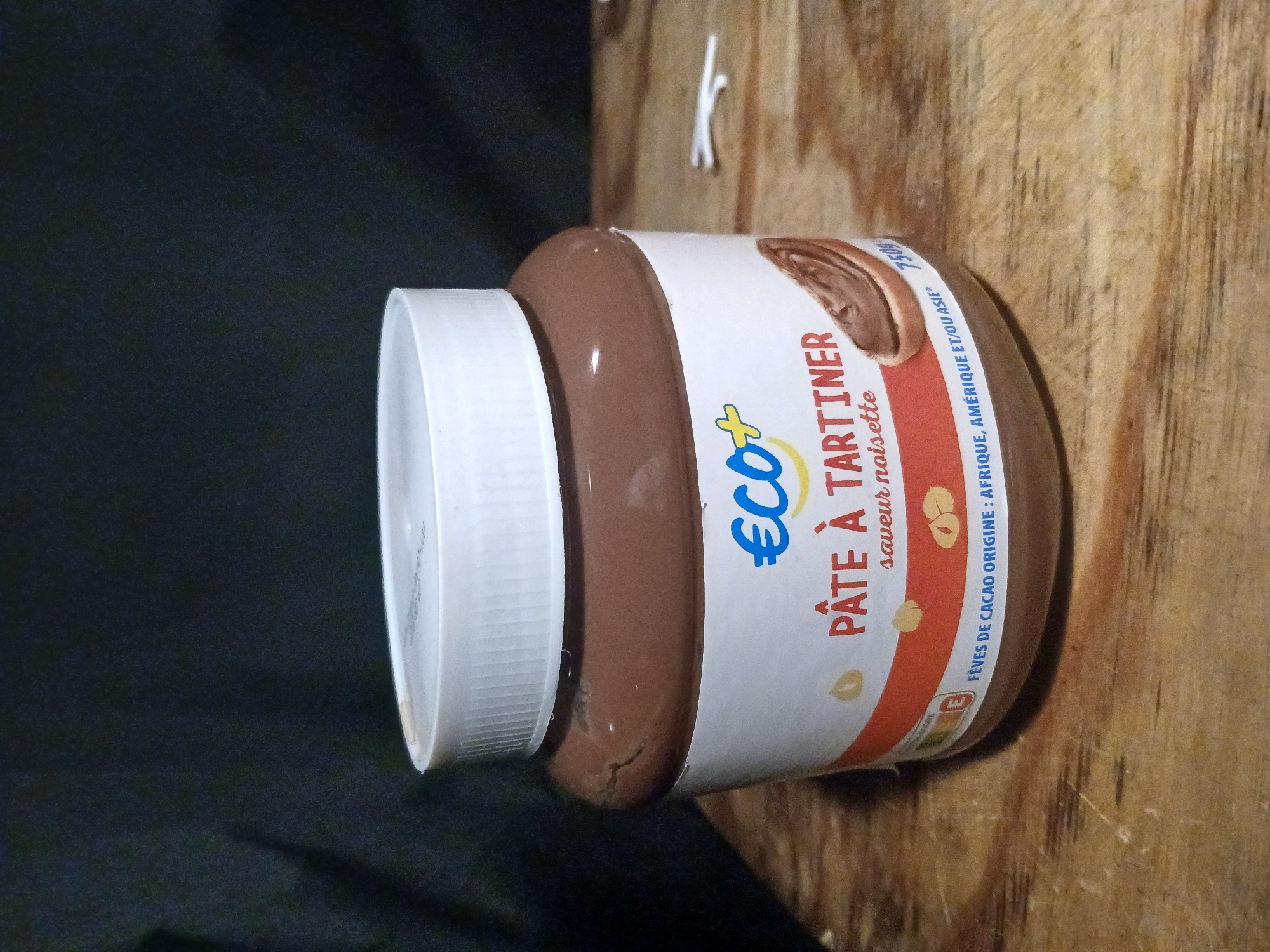 A jar of hazelnut spread, labeled "ECO+" in French and featuring an image of the spread on toast, is placed on a wooden surface. The label also states "PÂTE À TARTINER," meaning "spreadable paste." The jar is predominantly brown with a white lid. The background is dark and out of focus. The image does not contain any information about the city of Douai, France.