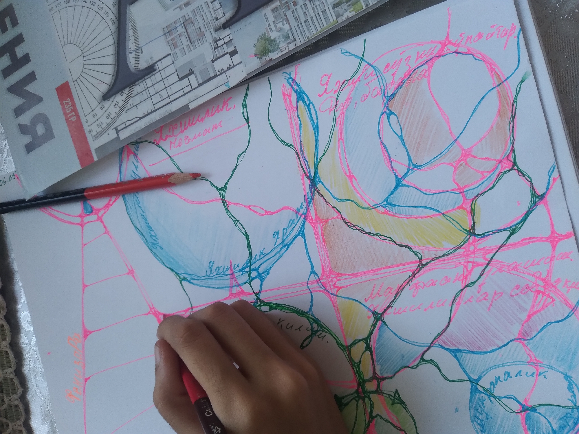 The image shows a close-up of a hand holding a red marker, drawing on a white sheet of paper covered in a network of colorful lines in pink, green, and blue. The drawing appears abstract, with circular shapes and interconnected lines. There is also a red pencil lying on the paper, and a ruler with architectural drawings and Cyrillic text is partially visible in the background. The lighting suggests an indoor setting. There is no visible indication of the city of Baki, Uzbekistan.