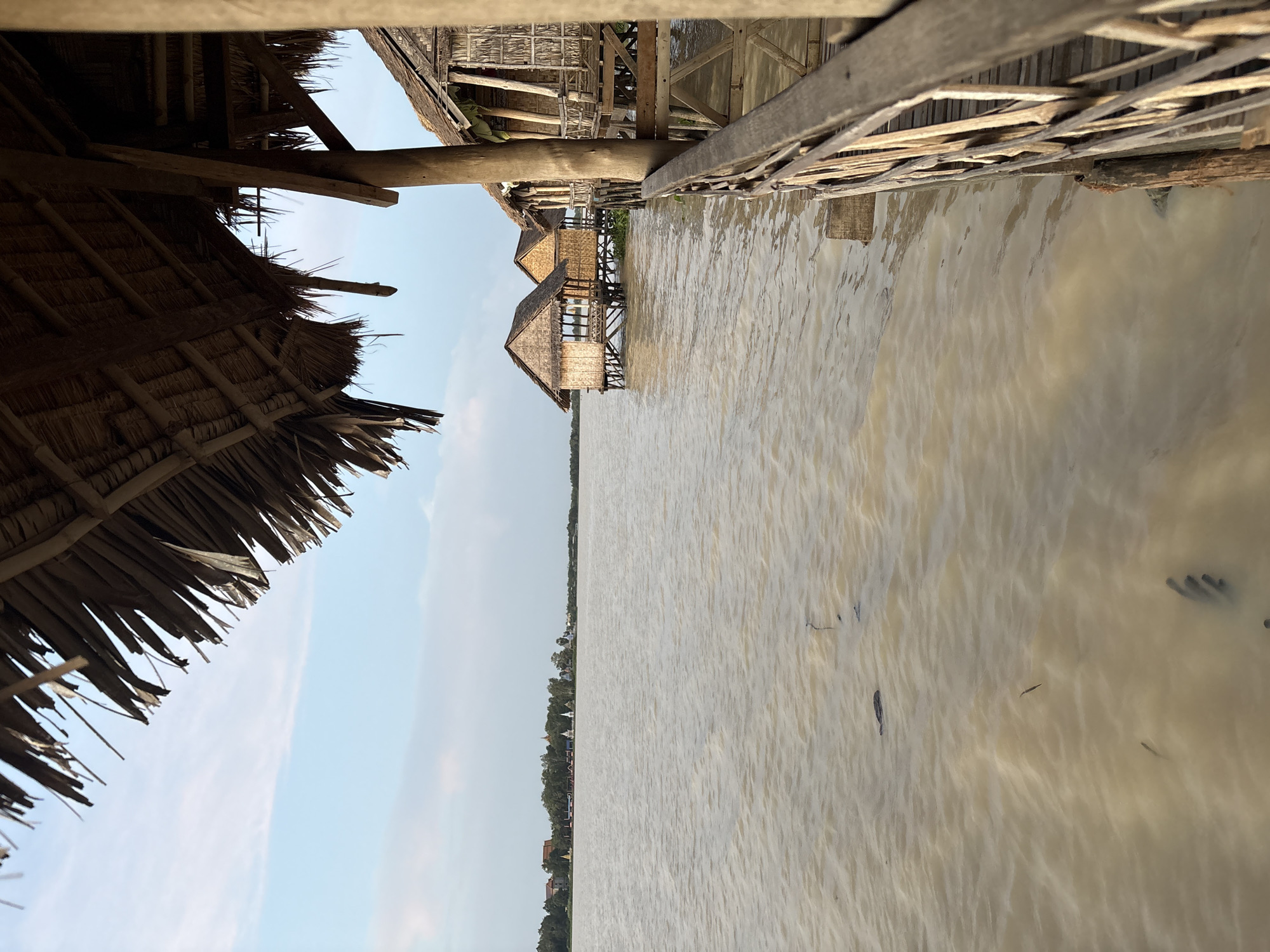 This image depicts a waterfront scene with stilt houses constructed from natural materials. The houses, with thatched roofs and woven walls, are situated over a murky, brown body of water. In the foreground, a wooden railing and walkway are visible, suggesting a visitor area. The water is calm with ripples, and some debris or aquatic plants are present on the surface. In the distance, a shoreline is lined with trees and buildings, including what appear to be white stupas, indicative of a temple or religious site. The sky is a pale blue with some wispy clouds. The overall impression is a tranquil, possibly rural or semi-rural setting in Tonle Bati, Cambodia.