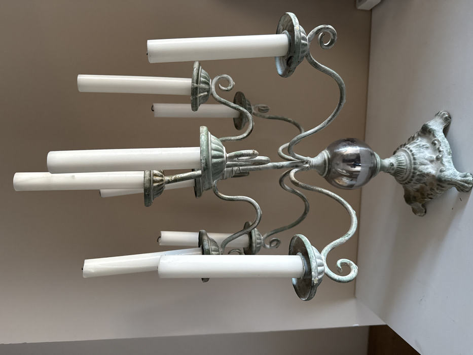 An antique-style metal candelabra stands centrally on a flat, light-colored surface, likely a table or shelf, against a plain, light beige wall. The candelabra exhibits a prominent light blue-green verdigris patina across its intricate design, suggesting age or an intentional aged finish. It features a decorative, four-footed base supporting a central stem, which incorporates a reflective, spherical, silver-toned element. Eight curved arms extend from the central stem, each terminating in a cup-shaped candle holder. Eight white, unlit, cylindrical taper candles are seated within these holders. The candles vary slightly in height, with some exhibiting minor dark marks at their wicks consistent with prior burning. No people or other significant objects are visible in the immediate vicinity. The scene depicts an indoor setting, purportedly within the city of Капитолий Кп, Russia, though visual cues within the image do not specifically identify this location.