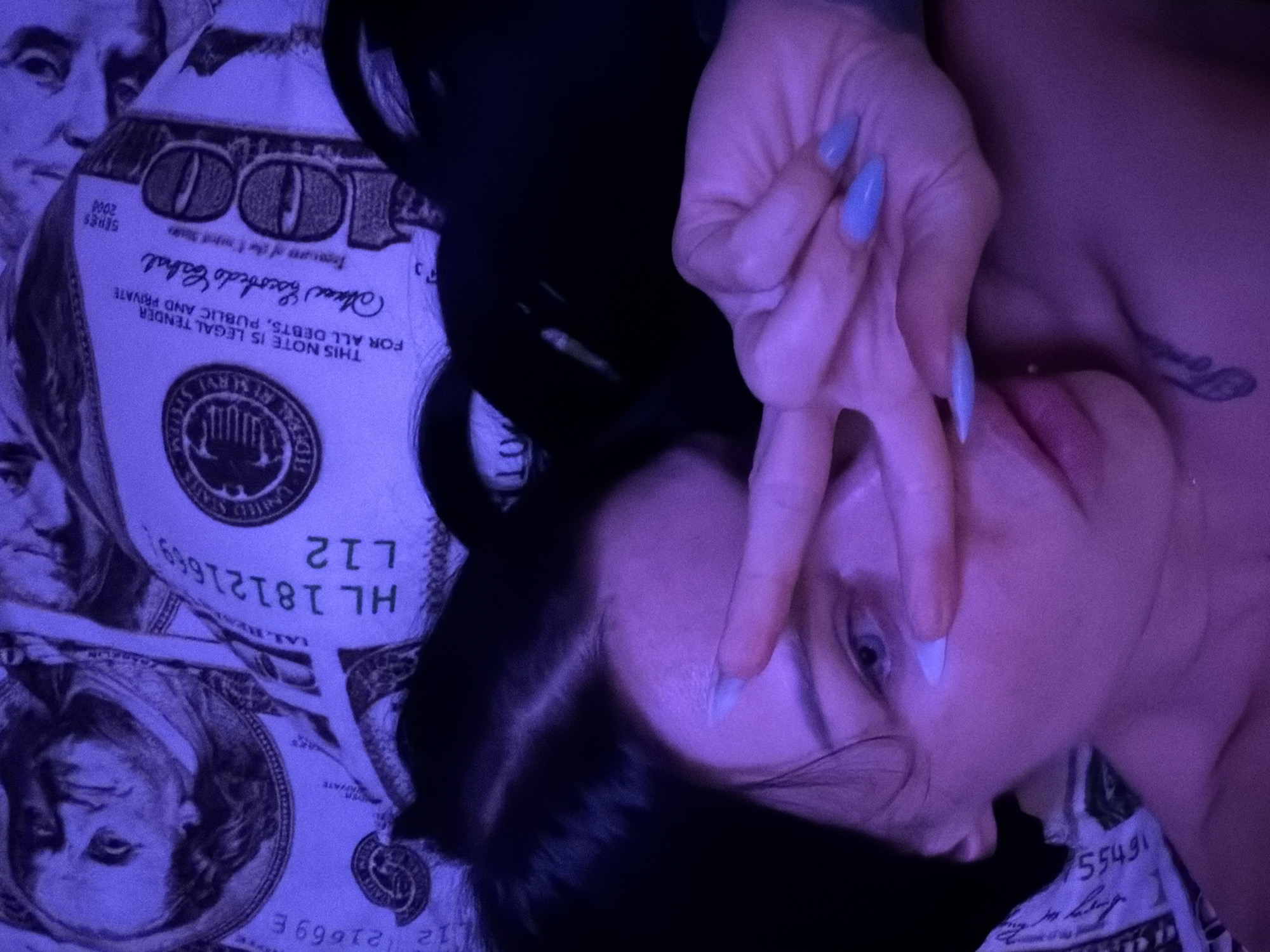 The image is a close-up selfie of a woman with dark hair. She is lying down, and the background appears to be a pattern of US $100 bills, illuminated by purple lighting. Her right hand is raised, with her index and middle fingers extended in a peace sign, partially obscuring her face. Her fingernails are painted a light blue color. She has a small tattoo visible on her collarbone. The image does not provide any information about the location in the city of Ufa, Russia.