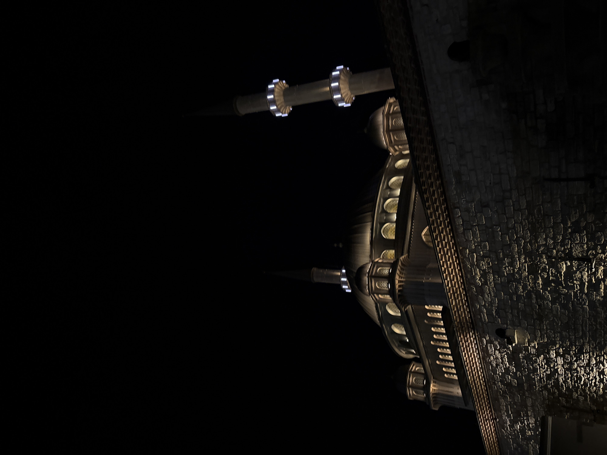 This forensic image captures the Taksim Mosque at night in Beyoğlu, Istanbul, Turkey. The central dome and adjacent smaller domes of the mosque are illuminated with warm, golden light, accentuating arched windows and intricate architectural elements. Two slender minarets are visible; one prominently on the right, and another partially in the background on the left. Each minaret features two balconies (şerefe) distinctively lit with bright white lights. In the foreground, a textured stone wall, topped by a decorative patterned eave, is illuminated from below by warm artificial uplights, creating deep shadows and highlighting the stone's rough surface. The background sky is completely black, indicating a nighttime scene. No individuals, vehicles, or other moving subjects are present.