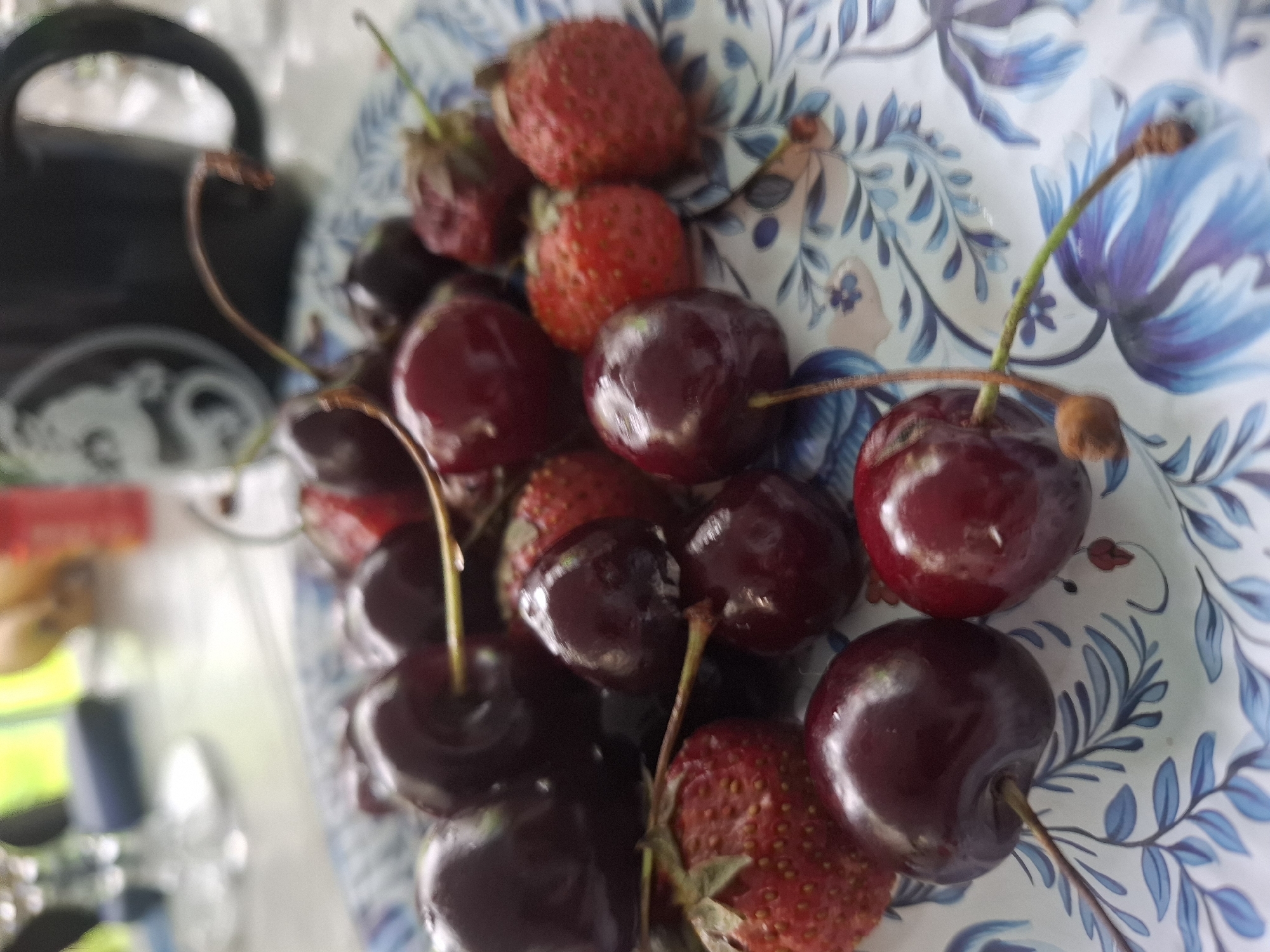 A close-up shot shows a plate filled with ripe cherries and strawberries. The plate has a blue and white floral pattern. In the background, out of focus, a dark mug with a white design and a glass are visible. The scene appears to be an outdoor setting, possibly a patio or garden, with greenery in the far background. The image does not provide any information about the city of Utulik, Russia.