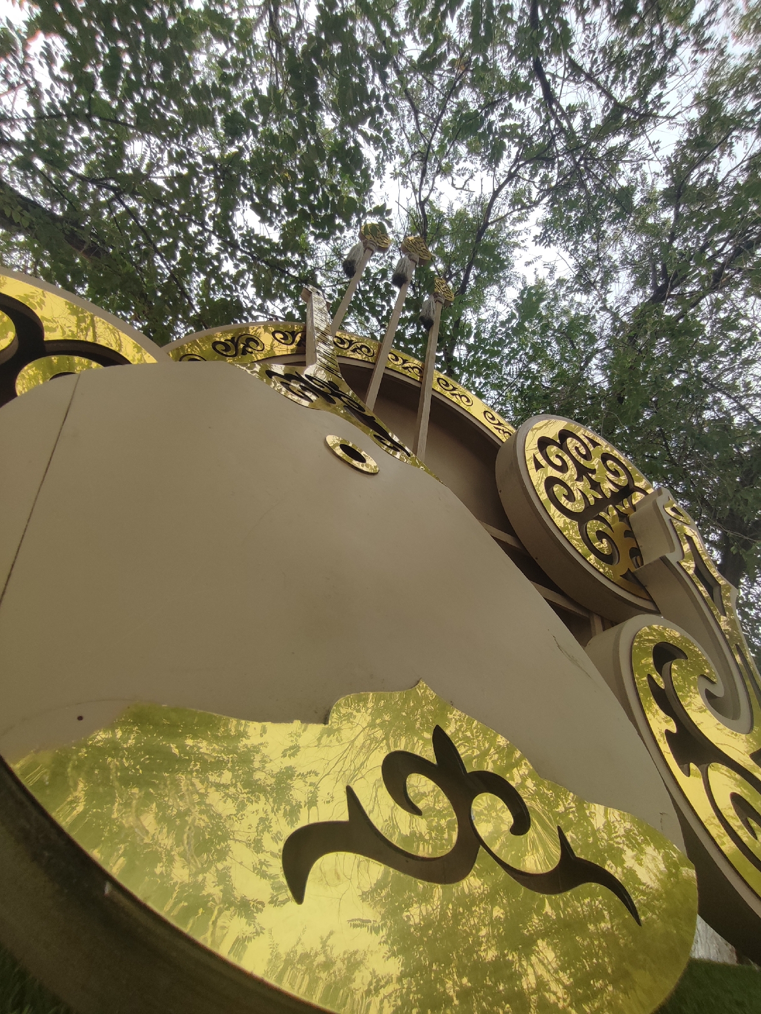 The image is a low-angle, close-up shot of an ornate, cream-colored sculpture with intricate gold metallic detailing. The sculpture features swirling patterns and decorative elements, and appears to be quite large, possibly part of a gate or monument. Above the sculpture, the branches and green leaves of trees are visible against a bright, overcast sky. Reflections of trees and the sky are visible on the gold surfaces of the sculpture. The sculpture is outdoors, with grass visible in the lower foreground. This sculpture is located in the city of Qonaev, Kazakhstan.