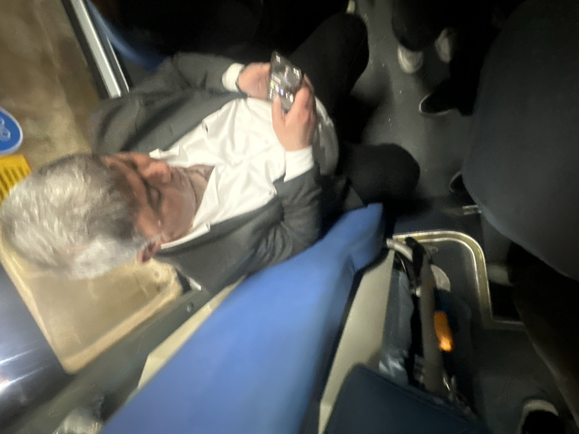 A person with gray hair, wearing a gray suit and white shirt, is sitting in a blue airplane seat. They are holding a smartphone in their hands. Other seated passengers are visible. The location is unknown.