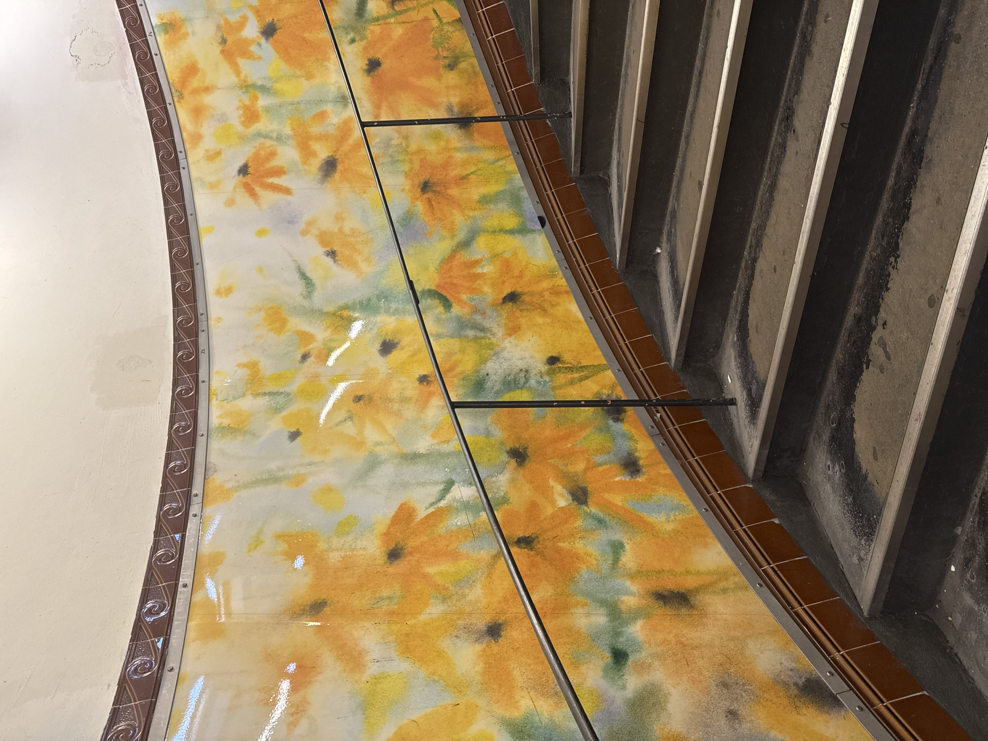 The image depicts a close-up view of a stairway landing, focusing on the juncture between the wall and the staircase. The wall features a decorative border near the ceiling with a patterned design. Below this border, the wall is painted in a neutral color. Adjacent to the wall is a section of the landing decorated with a mural of yellow and orange floral patterns. This mural is segmented into panels, held together by metal supports. The staircase is made of concrete with treads and risers. The edges of the steps are visible, and the stairs appear worn with some discoloration. The location and time of day are unknown.