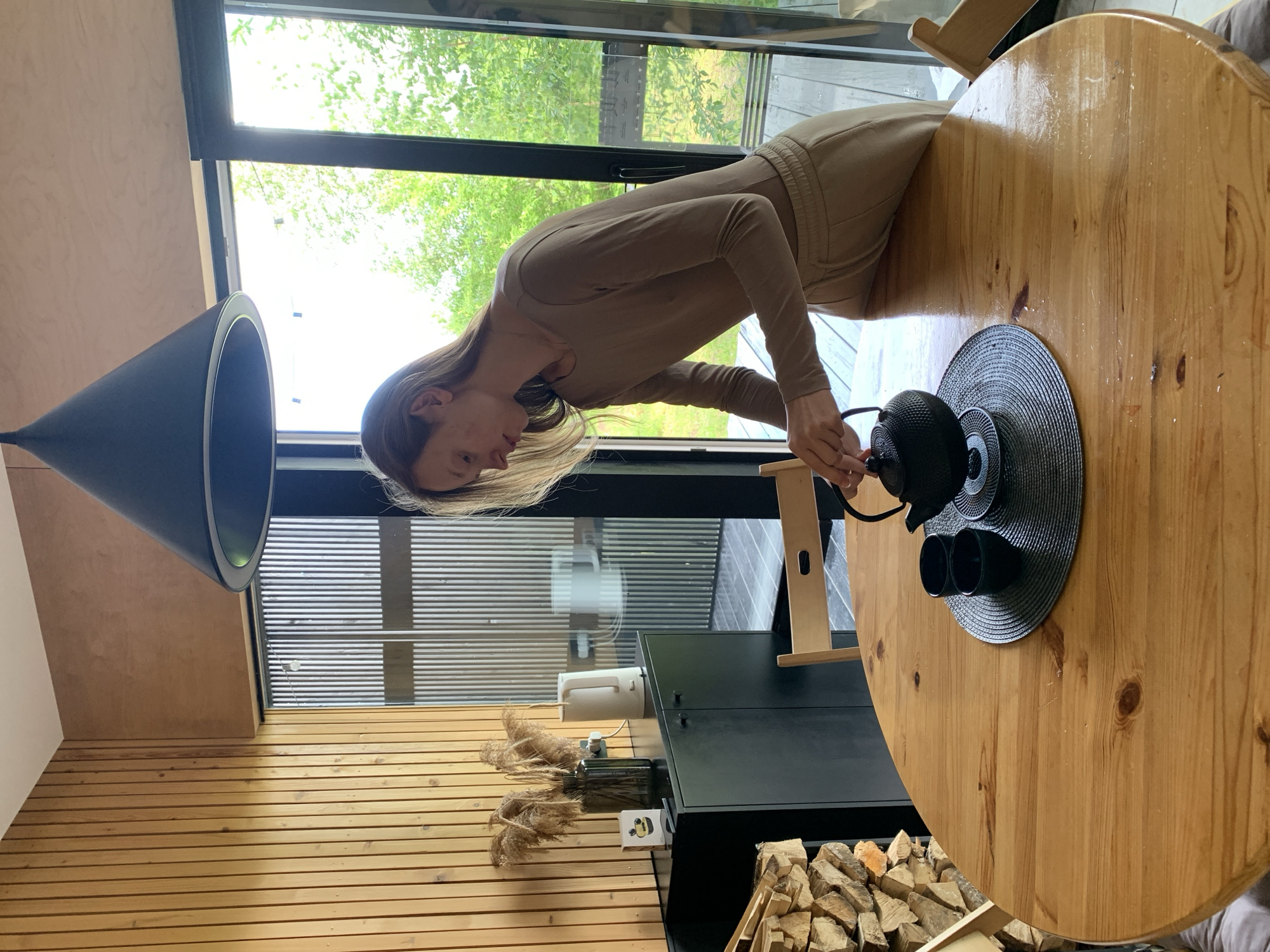 In a rustic interior with wood-paneled walls, a young woman with long, light brown hair, wearing a tan long-sleeved top and matching pants, is pouring tea. She is leaning over a round wooden table where a black teapot and two small black cups are arranged on a dark grey placemat. The room features a dark cabinet with a white electric kettle and dried reeds in a vase on top. To the left, a stack of firewood is visible. A large black pendant light hangs from the ceiling. Through a glass sliding door, lush green trees and vegetation are visible, suggesting an outdoor setting beyond. The scene appears to be in a dwelling, possibly a home, within the vicinity of Мяжанскі сельскі Савет, Belarus, characterized by its natural and somewhat secluded environment.