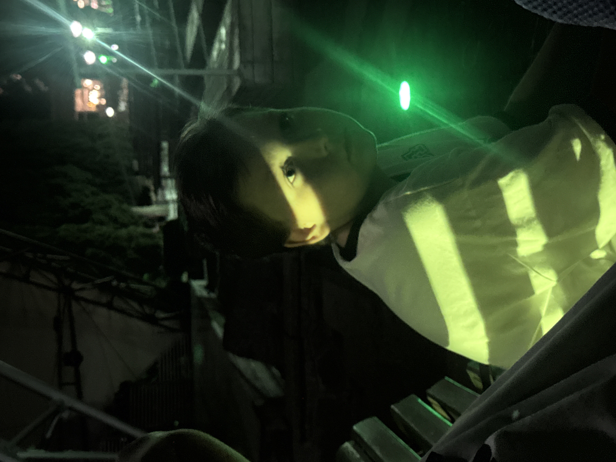 In an outdoor nighttime setting, possibly in the city of Yalta, Ukraine, a young boy sits on a bench. He is illuminated by a strong, bright green light that casts sharp shadows across his face and white t-shirt. The boy is looking towards the right side of the frame, his expression unreadable due to the lighting. Behind him, the background is dark and out of focus, with hints of distant lights and trees suggesting an urban or park environment. The overall impression is one of artificial illumination creating a dramatic and somewhat surreal atmosphere.