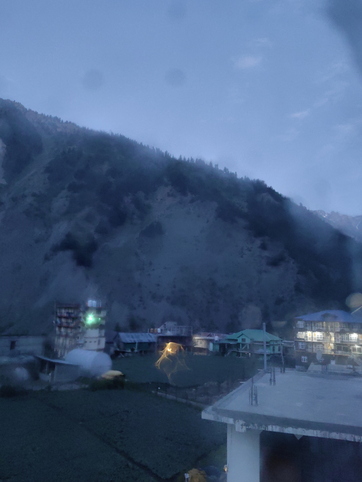 This is a photograph of a mountainous landscape with a village nestled at the foot of a slope. The image appears to have been taken at dusk or during a period of low light, as indicated by the muted colors and the artificial lights emanating from some buildings. The sky is overcast, and there is a noticeable mist or fog obscuring the upper slopes of the mountain, which is covered in sparse vegetation and trees.
In the foreground, there is a dark, cultivated field, possibly farmland. Several buildings are visible in the midground. One prominent structure is a multi-story building with illuminated windows, suggesting it is a hotel or residential building. Next to it, there's a building with a green roof. To the left, another tall building with several floors and lit windows is visible. Smaller structures and what appears to be a greenhouse or a covered farm area are also present.
The overall scene depicts a rural or semi-rural settlement in a mountainous region. The lack of distinct human figures or specific cultural landmarks makes it difficult to pinpoint a precise location without additional context. This image is not of Udaipur, India, as Udaipur is located in a desert region and is known for its lakes and palaces, not mountainous terrain and mist.