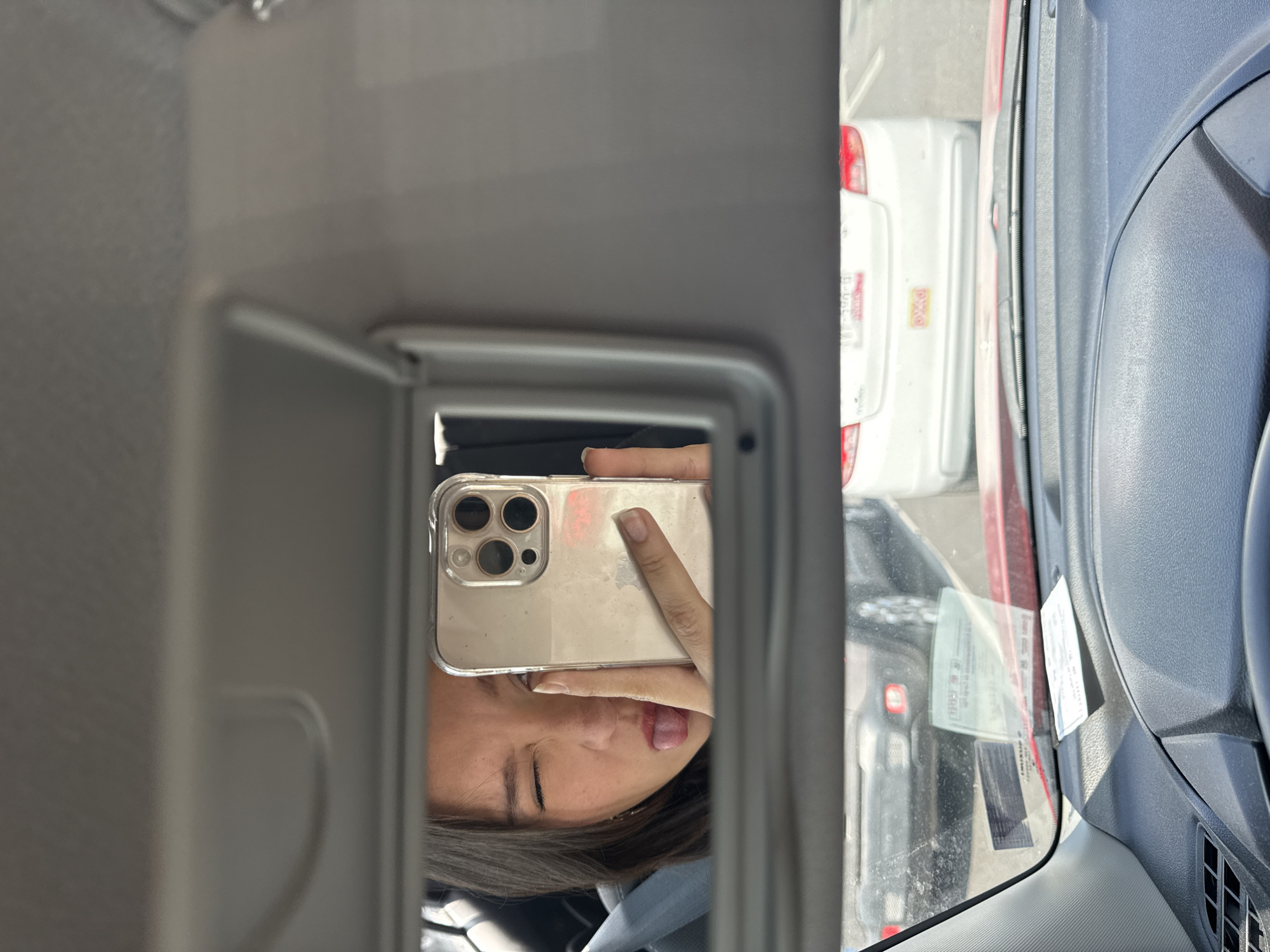 A young woman takes a selfie in a car mirror with her tongue sticking out. The car appears to be in Puebla, Mexico, based on the license plates of the vehicles visible outside the car window. Other cars are parked close to the subject's car. The car mirror, the subject's face and hand holding the smartphone, and exterior parked cars are the main subjects. The woman is not interacting with anything but her phone and herself.