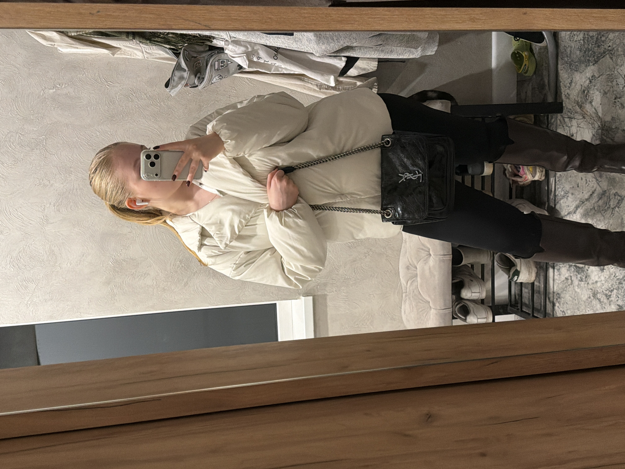 A woman stands in what appears to be a room in a residence, using her phone to take a mirror selfie. She is wearing a light-colored puffer jacket, black leggings, and brown knee-high boots. She is also wearing earbuds. A black handbag with a silver chain hangs from her shoulder. Behind her, a shoe rack is visible, holding several pairs of shoes. A clothes rack with hanging garments is also visible. The photo is taken in Rossiyskiy, Russia.