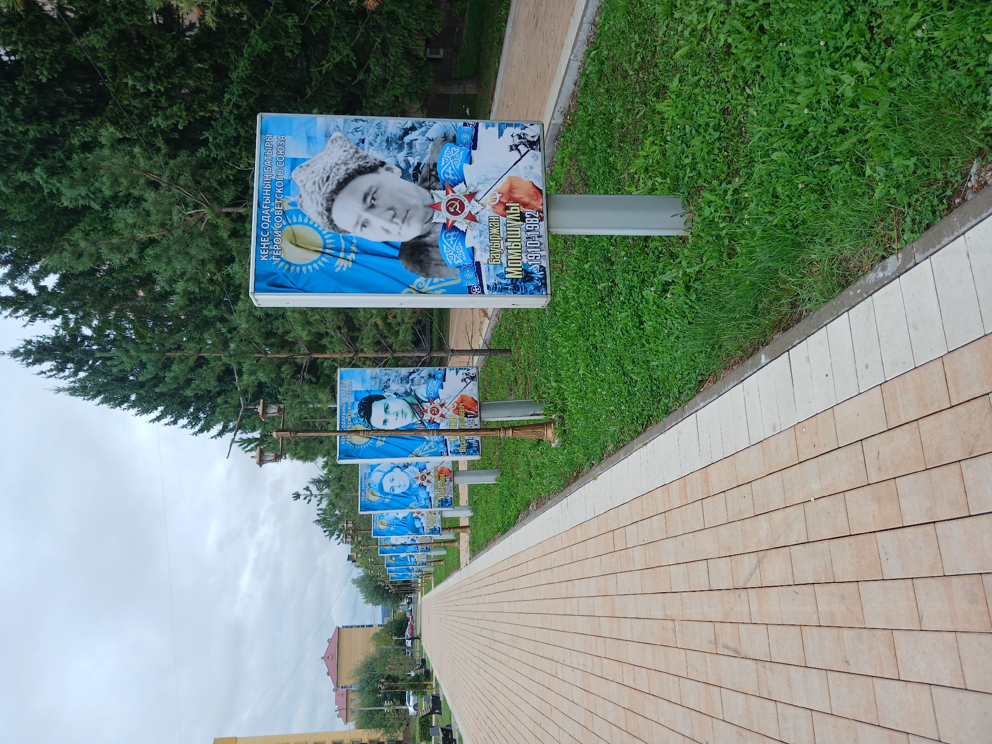 A paved walkway lines the left side of the image, bordered by a strip of grass on the right. Along the grass, a series of tall, rectangular billboards are erected on poles, each displaying a portrait of a person against a blue background with the Kazakh flag. The billboards appear to be in honor of heroes from the Soviet Union. Trees line the far right of the image. In the background, there are buildings, more trees, and a cloudy sky. The scene is located in the city of Shchuchinsk, Kazakhstan.