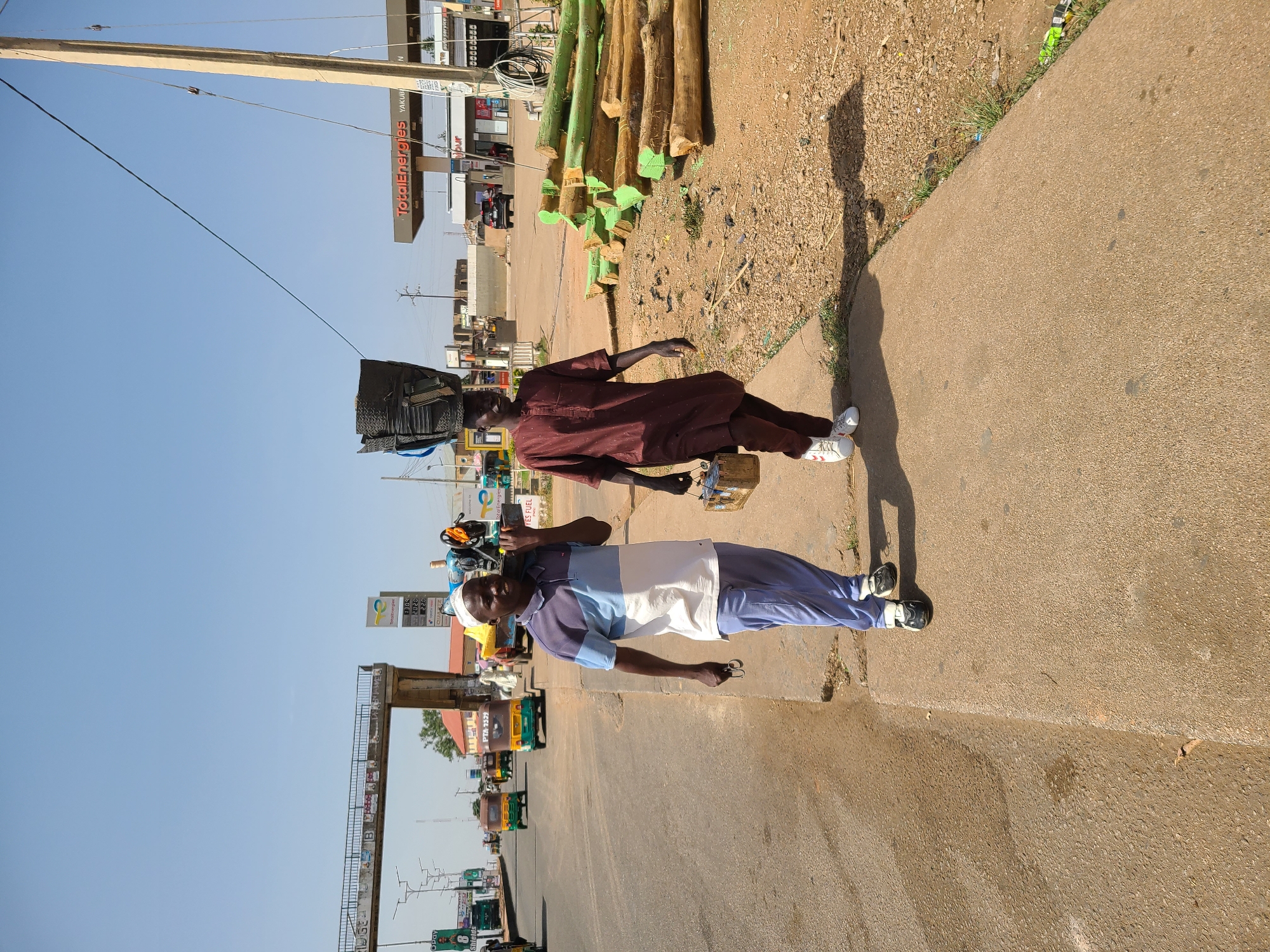 The image depicts a street scene in Jos, Nigeria. Two men are walking on a sidewalk beside a road with traffic. One man carries a heavy load on his head, while the other holds small items. The background includes a TotalEnergies gas station, a pedestrian bridge, piles of logs, and commercial buildings. The scene is well-lit by sunlight.