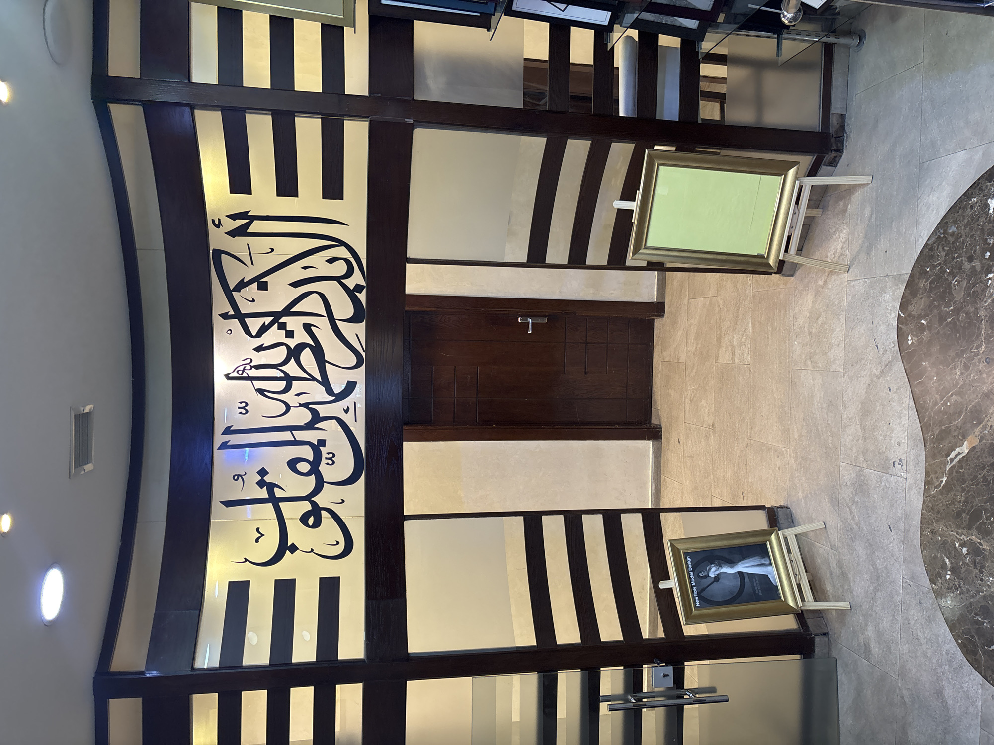 The image shows the interior of a shop with Arabic calligraphy on the wall. In the foreground, there are two easels displaying framed pictures. The easel on the left displays a framed picture with text that reads "New Italy Model Design" and a black and white image of a woman in a dress. The easel on the right displays a framed picture with a blank green background. In the background, there is a wooden door. The shop appears to be located in Amman, Jordan.
