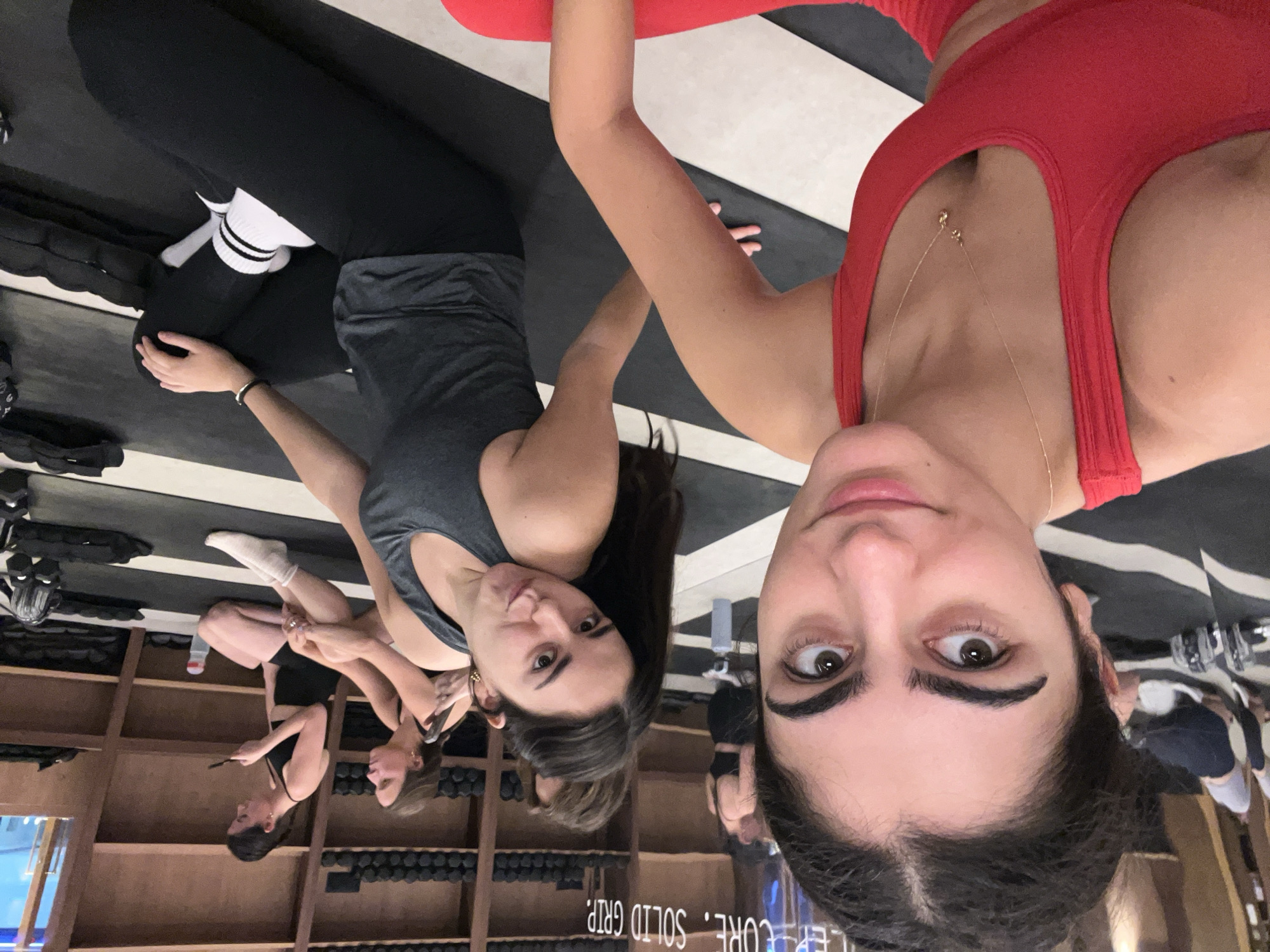 The media file shows a group of young women practicing yoga indoors. The setting appears to be a studio with wooden shelves filled with weights in the background. The women are dressed in athletic wear, with some wearing leggings and sports bras. One woman in the foreground is wearing a red sports bra and has a gold necklace. Another woman is wearing a grey tank top and black leggings with white striped socks. The floor has a black and white striped pattern. The image is taken from a low angle, with the subjects appearing upside down. The lighting suggests it is daytime. The overall mood seems casual and friendly, with some of the women smiling. There is text visible on the shelves in the background which reads "CORE. SOLID GRIP."