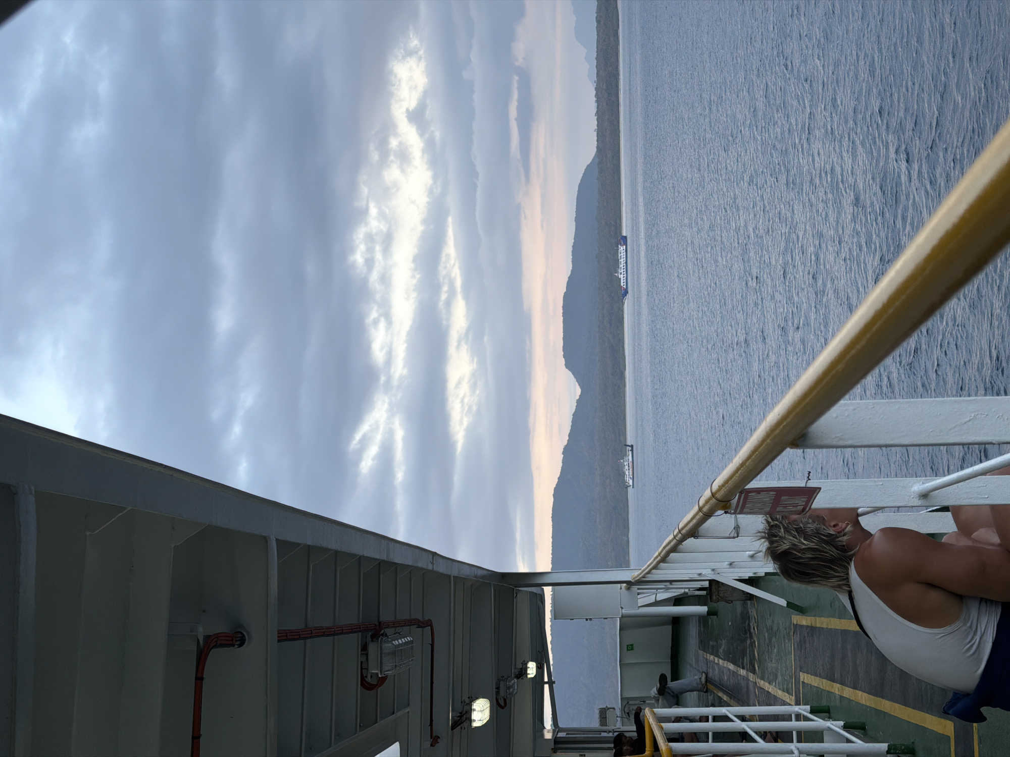 This is a medium shot of a person on the deck of a ferry, looking out at the sea. The person is wearing a white tank top and has blonde hair. They are sitting on the deck and leaning against the railing. In the background, there is a body of water and a distant island. A ferry boat is visible in the distance. The sky is cloudy and overcast. The location is Melaya, Indonesia.
