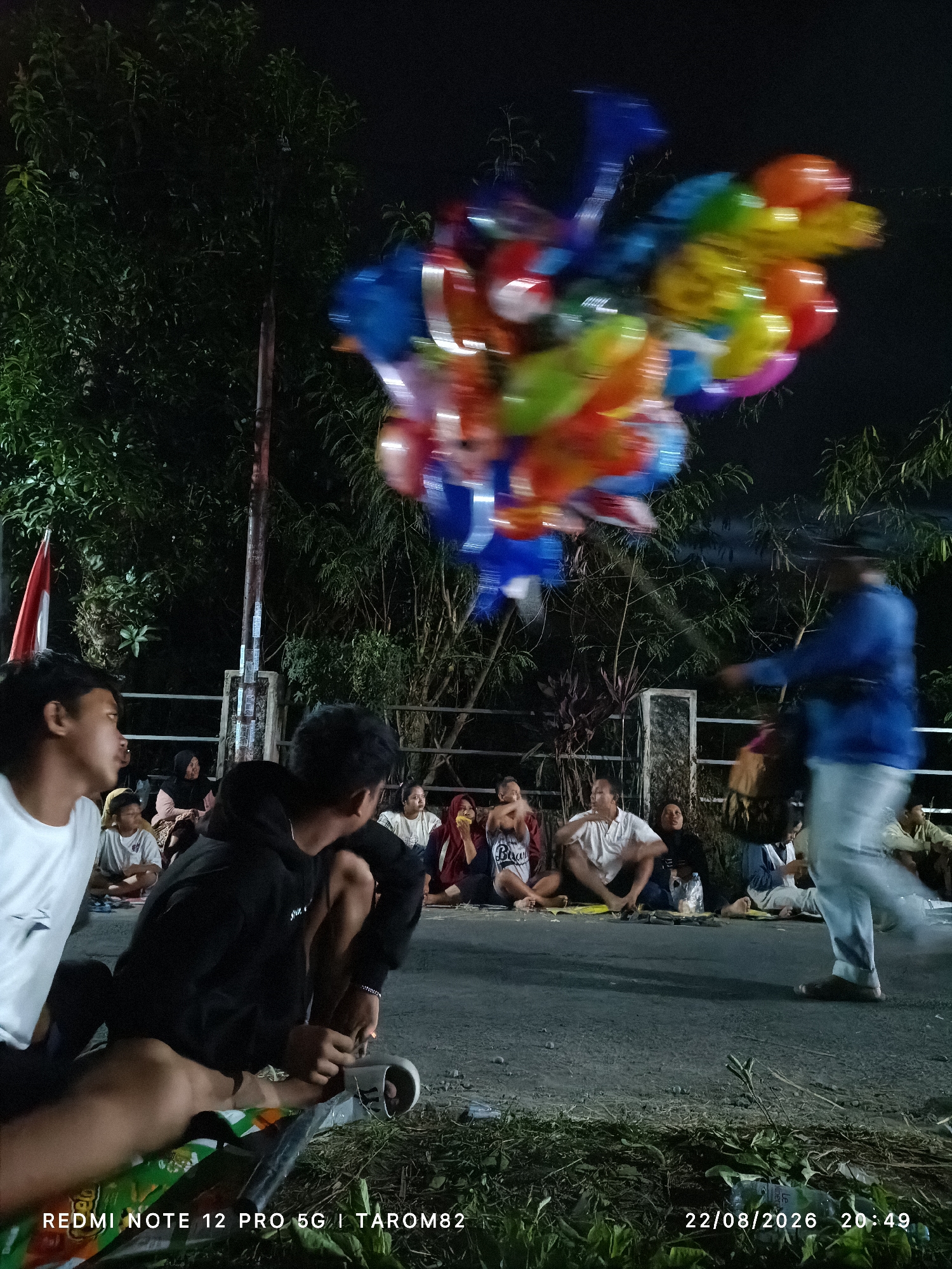 In Mundusewu, Indonesia, at night, a group of people are seated on the ground by the side of a road. In the foreground, two young men are sitting on the ground. One is wearing a white t-shirt and looking to the left, while the other is wearing a black hoodie and looking down. Behind them, several other people are seated on the road, some of whom appear to be families. A person in a blue shirt is walking away from the viewer on the right side of the image, carrying a bag. Above, a bunch of colorful balloons are blurred, suggesting they are in motion or the photo is taken with a slow shutter speed. The background consists of dark trees and foliage. The timestamp "22/08/2026 20:49" is visible in the lower right corner, along with "REDMI NOTE 12 PRO 5G | TAROM82" in the lower left corner, indicating the device and possibly the location of the photo.