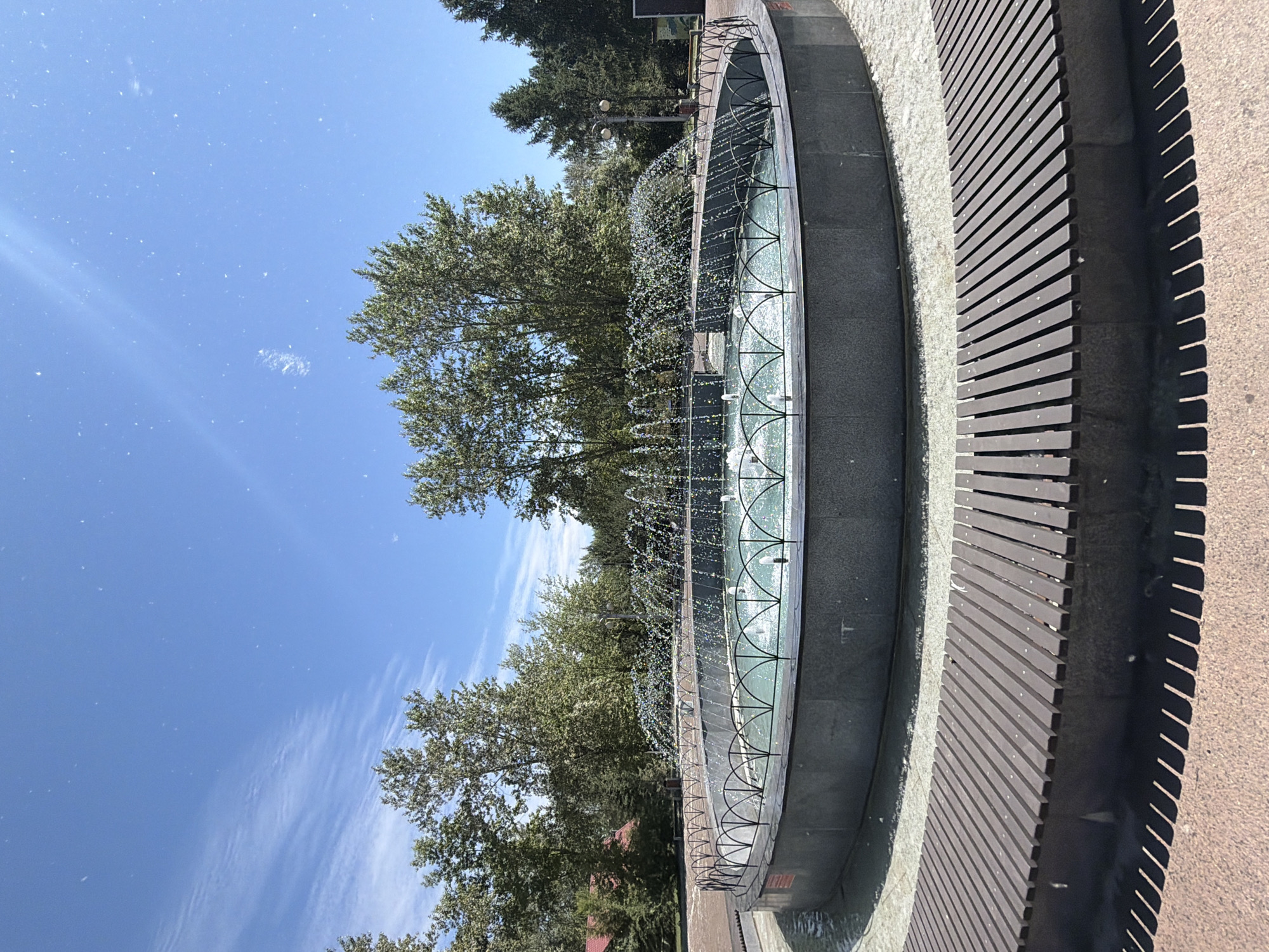 The image shows a large circular fountain in a park. The fountain has multiple jets of water spraying upwards, creating a shimmering effect. The fountain is surrounded by a grey stone base and a decorative metal fence. Beyond the fountain, there are several large trees with green foliage and a clear blue sky with some wispy white clouds. In the foreground, there is a curved wooden bench with metal supports, casting long shadows on the reddish-brown paved ground. The scene appears to be a public park or square. This is the fountain in Teatralnaya Square in Krasnoyarsk, Russia.