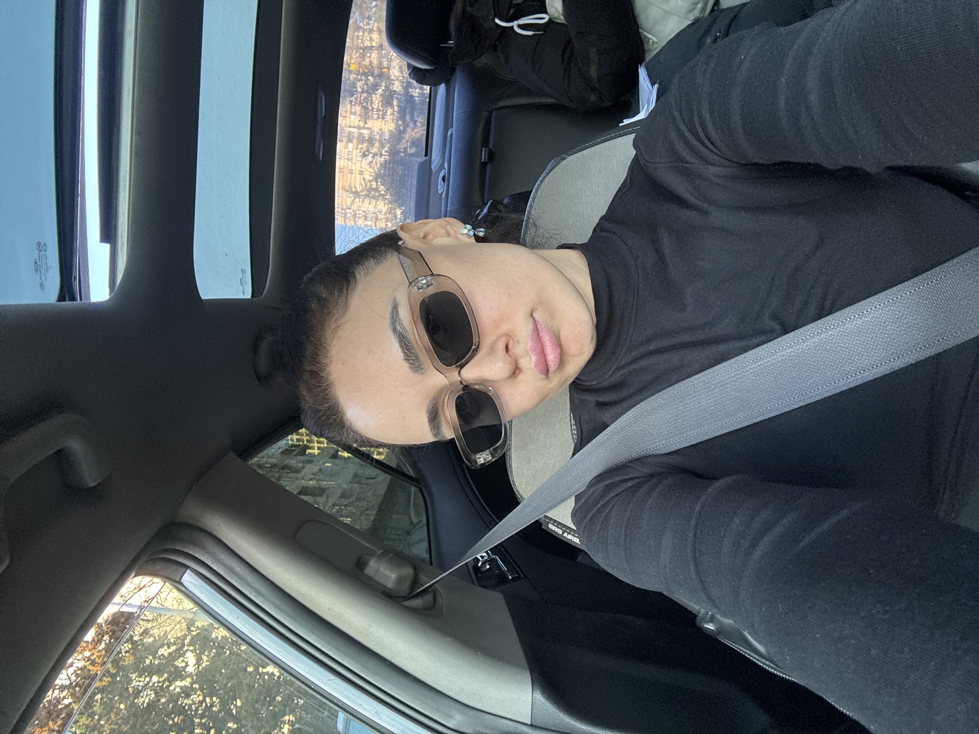 A female individual is seated in the front passenger or driver's seat of a vehicle, looking directly at the camera. She wears a black long-sleeved turtleneck shirt, rectangular light brown-tinted sunglasses, and small, light-colored earrings. Her hair is pulled back, and her lips are slightly pursed. A grey seatbelt is secured across her chest and lap. The vehicle's interior includes a sunroof and two-tone grey and beige upholstery on the seats. Through the left-side window, bare trees are visible, illuminated by sunlight. In the background, visible through the rear window and sunroof, are multiple multi-story residential buildings with numerous windows, consistent with the urban architecture of apartment blocks found in Tbilisi, Georgia. A dark jacket is visible on the rear seat. The sky is clear.