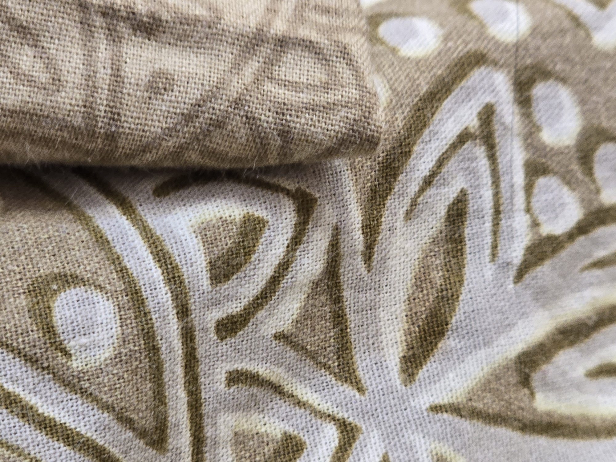 The image is a close-up, macro shot of a textile with a patterned design. The fabric appears to be a natural fiber, possibly linen or cotton, with a visible weave texture. The pattern consists of organic, floral or mandala-like shapes in shades of brown and white, printed on a lighter, off-white background. The fabric is folded, creating different layers and shadows that add depth to the image. The lighting is soft and diffused, highlighting the texture of the material and the details of the print. There is no discernible location in the city of Ulan-Ude, Russia, nor are there any people, actions, or interactions visible in the image. The scene is purely focused on the fabric and its pattern.