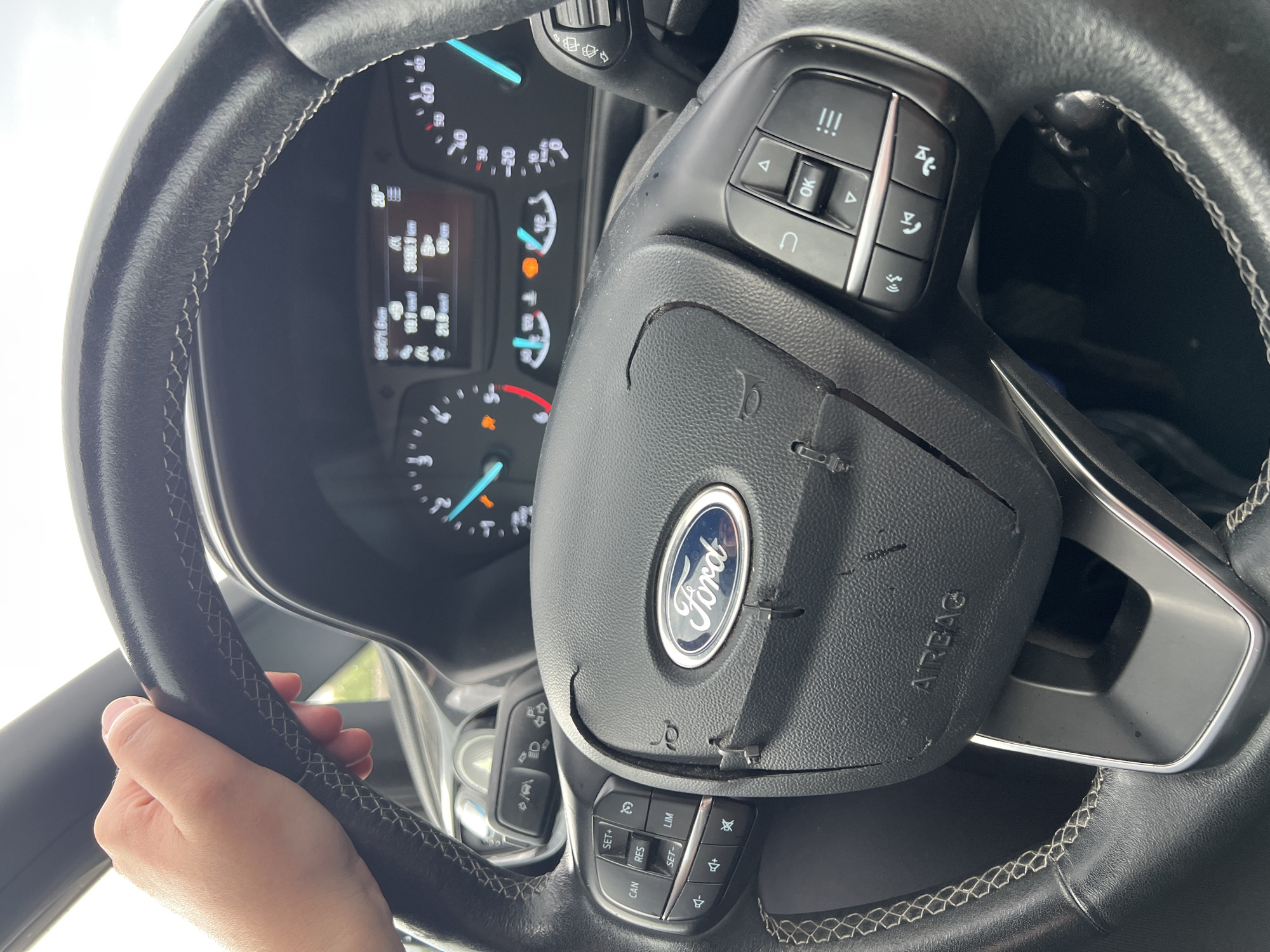 A person's hand grips a black leather steering wheel inside a vehicle. The steering wheel has visible damage, with the leather peeling away in several places. The Ford logo is prominent in the center of the steering wheel. The dashboard displays various indicators and a digital screen showing speed, distance, and temperature. The vehicle appears to be parked or stationary. The overall scene suggests a close-up view of a damaged car interior, likely in a location such as Molfetta, Italy, though this cannot be definitively confirmed by the image alone.
