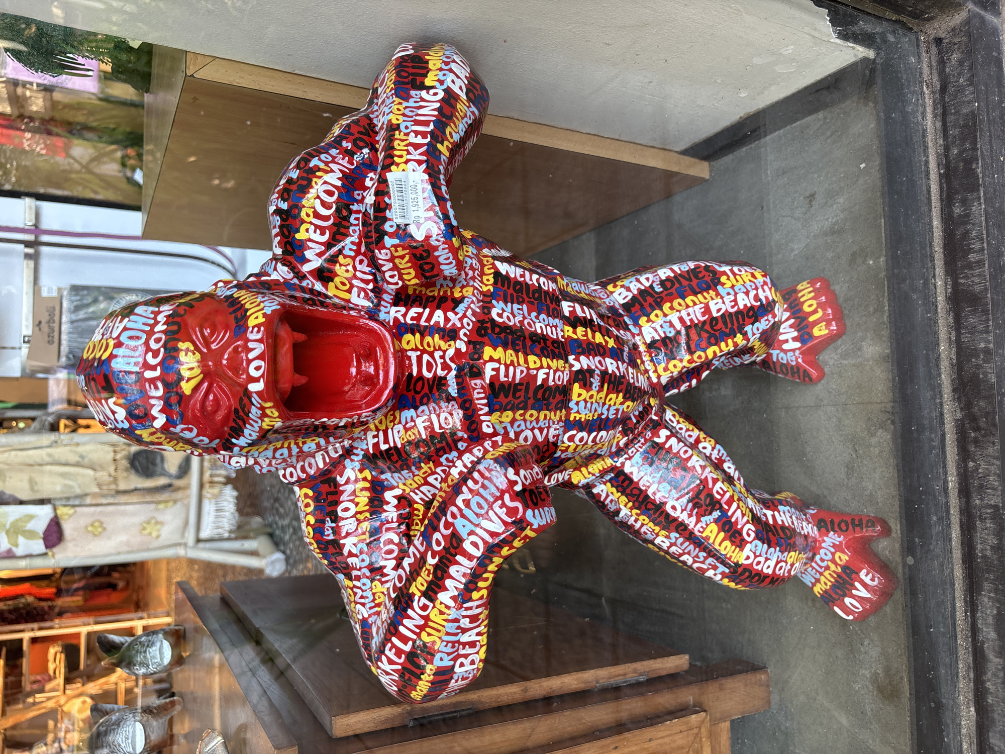 A vibrantly painted, life-size statue of a gorilla stands prominently in a shop window in Kuta Selatan, Indonesia. The gorilla is covered in a collage of words and phrases in various colors, including "Welcome," "Aloha," "Love," and beach-themed terms like "Beach," "Surfing," and "Snorkeling." The statue has a bright red face with its mouth open in a roar, revealing sharp teeth. It is positioned in a dynamic stance, with its arms outstretched. Behind the gorilla, the interior of the shop is visible, with displays of clothing and merchandise on shelves and a ladder. The scene is viewed through a glass window, with reflections and the surrounding environment of a street or walkway visible on the glass surface. The overall impression is a commercial display designed to attract attention.