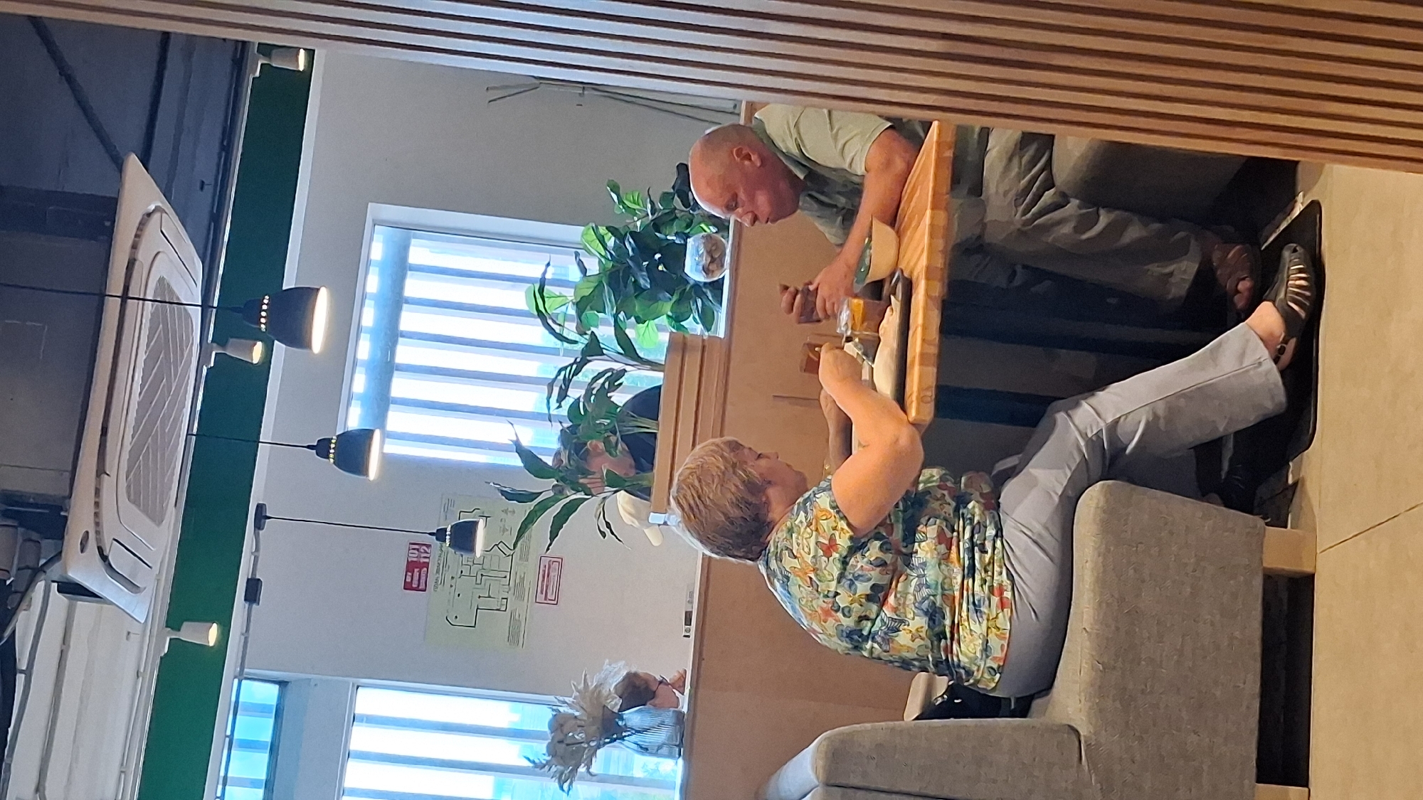 Two individuals, a man and a woman, are seated at a wooden table in a cafe in Novosibirsk, Russia. The woman, with short blonde hair, wears a colorful floral-patterned shirt and light blue pants. She is actively eating, holding a fork in her right hand. The man, bald and wearing a green shirt, is also eating and appears to be talking, with his mouth open. A white bowl, a glass, and other food items are on the table. The cafe has large windows with vertical blinds, potted plants, and overhead lighting fixtures. The overall ambiance suggests a casual dining experience.