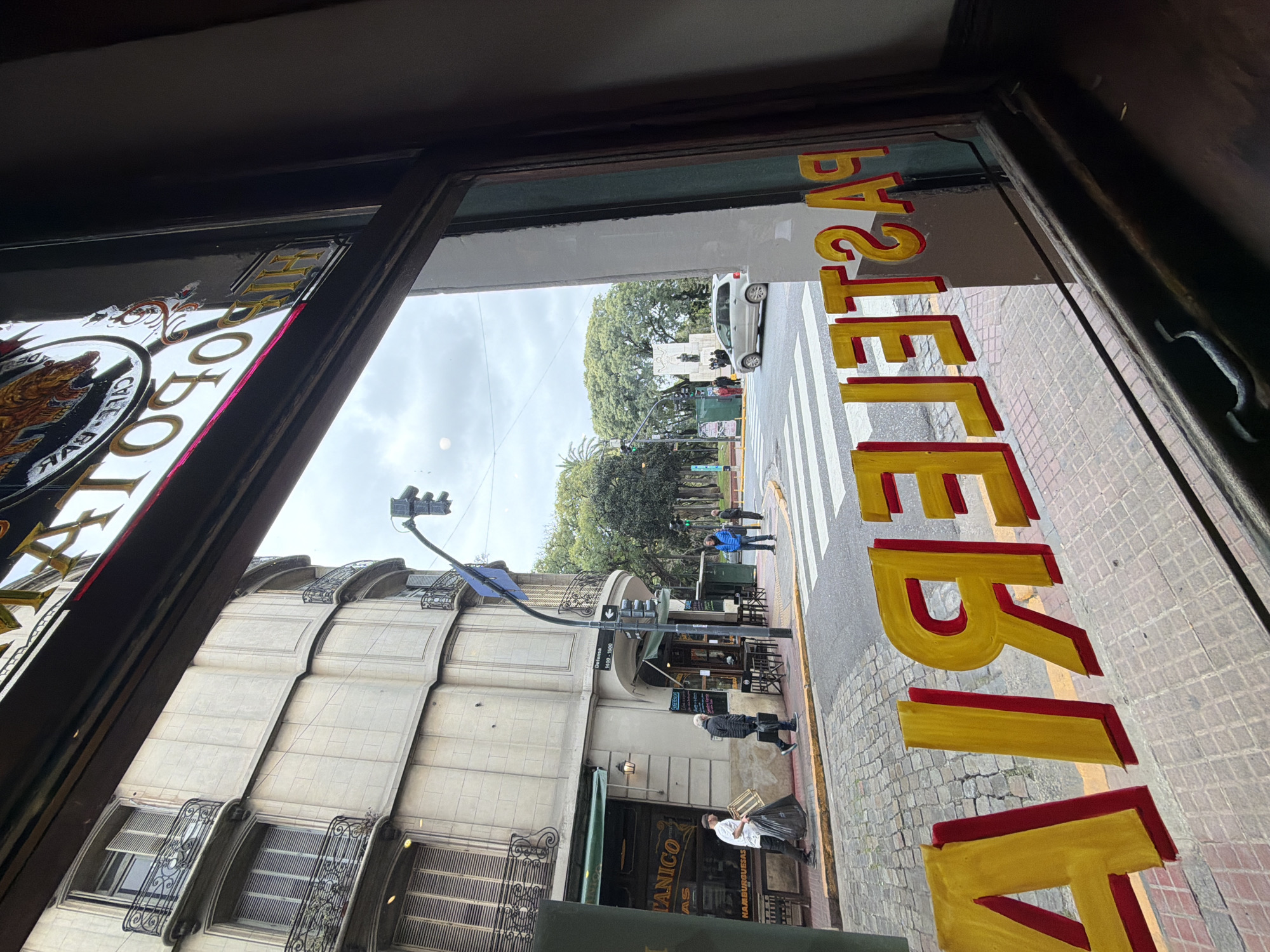 View from inside a shop window with the word "Pasteleria" painted on it. Outside, there is a street in Buenos Aires, Argentina, with a crosswalk, traffic lights, a monument, buildings, and people walking on the sidewalk. One shop is named "Botanico." The sky is overcast.