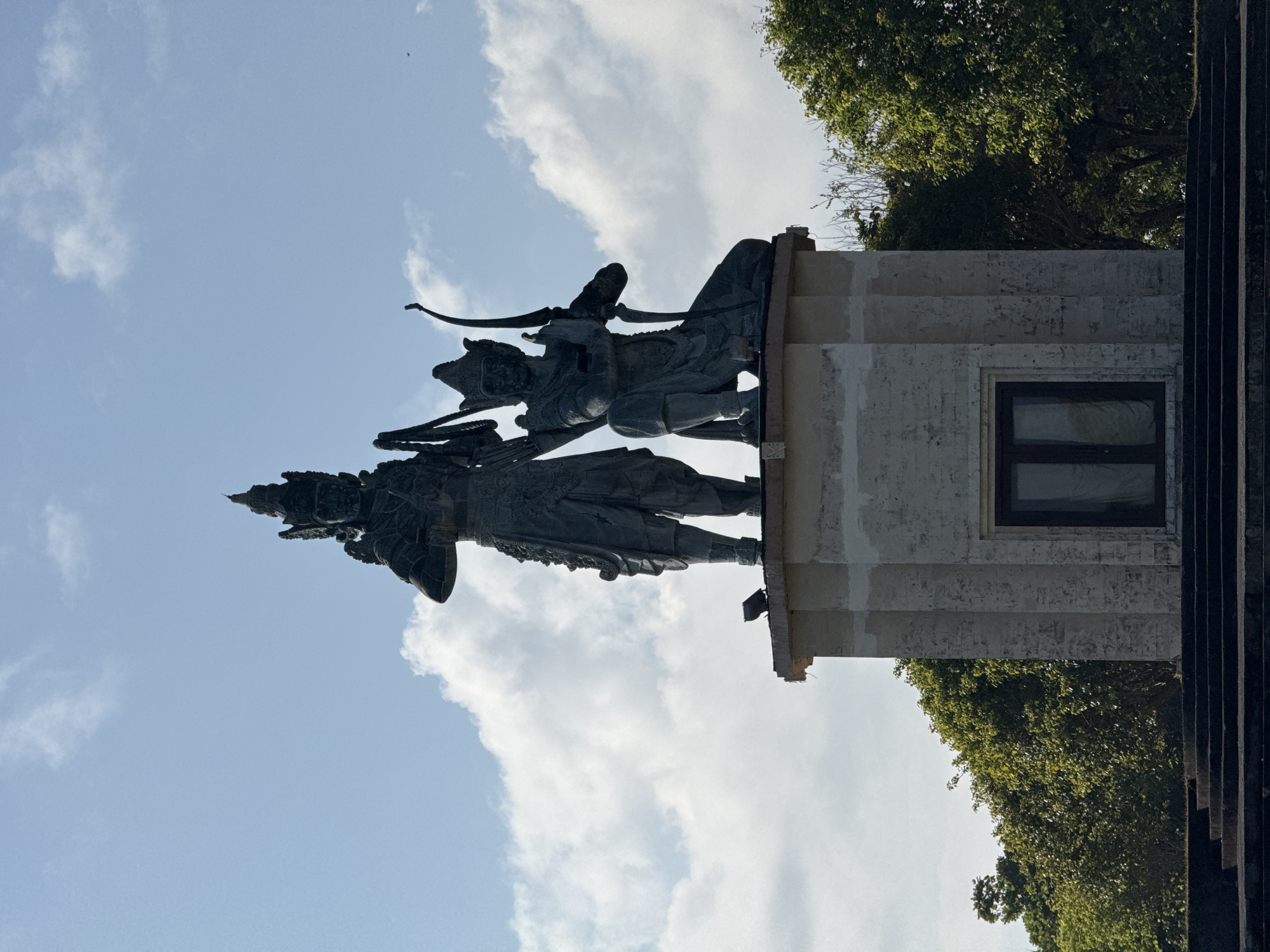 In Kuta Selatan, Indonesia, a large bronze statue depicting two figures stands atop a rectangular, stone pedestal with a dark-framed window. The figure on the left is standing and holds a rope or chain. The figure on the right is kneeling, holding a bow with an arrow drawn. Both figures wear ornate headdresses and traditional attire. The pedestal is situated on a tiered base. Lush green trees surround the base of the pedestal, and a bright blue sky with scattered clouds forms the background. A small bird is perched on the pedestal near the standing figure, and a tiny dark object, possibly a bird, is visible in the sky on the far right.