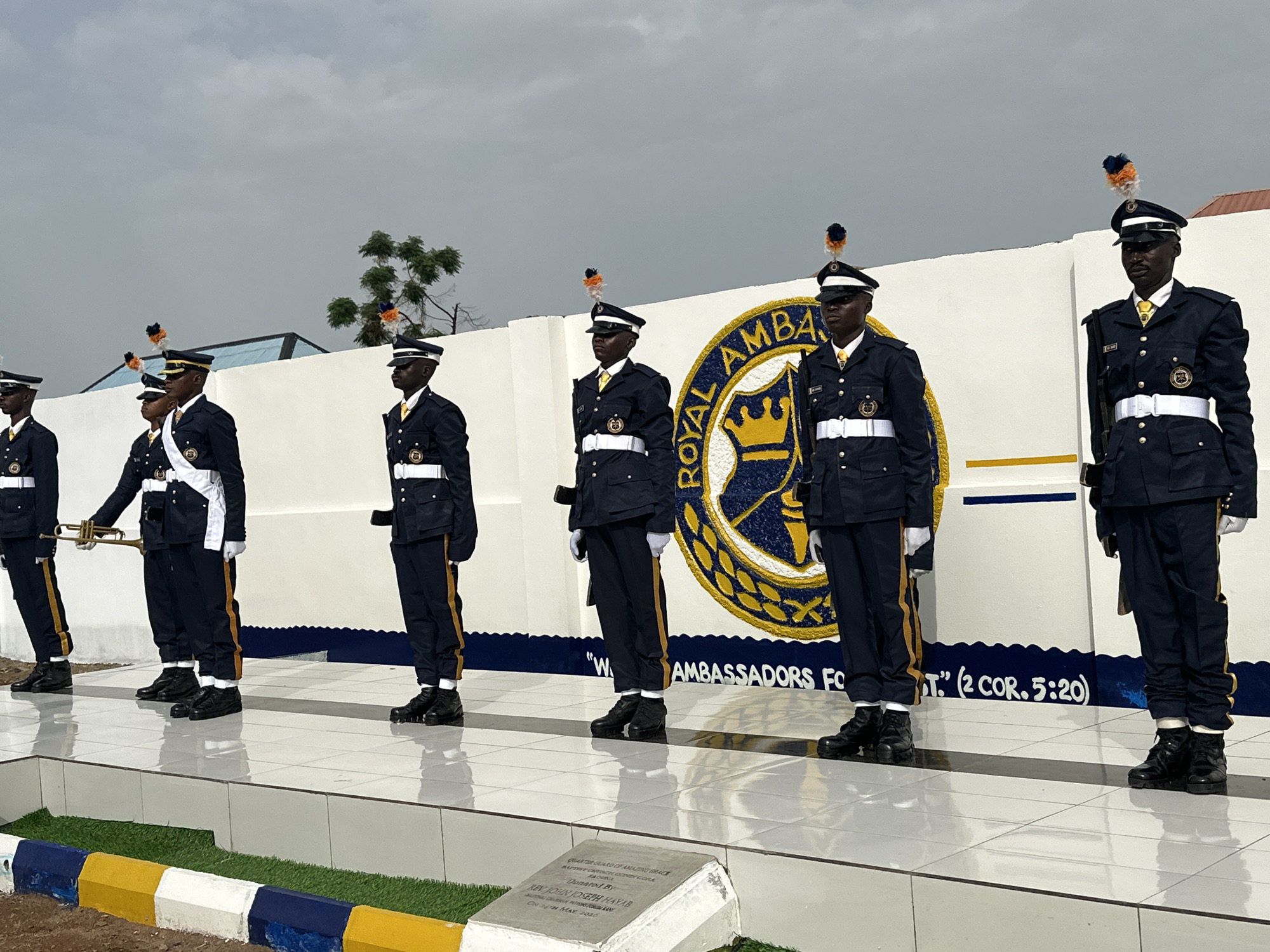 A photograph captures a group of six individuals, all Black men, standing at attention in a line on a raised platform with a white tiled surface. They are dressed in dark blue military-style uniforms with white belts and hats adorned with orange and black plumes. The second individual from the left holds a golden trumpet. Behind them is a white wall featuring a blue and gold emblem with the words "ROYAL AMBASSADORS FOR CHRIST." Below the emblem, script reads, "We are Ambassadors for Christ." (2 COR. 5:20)."
At the bottom left corner of the image, a commemorative plaque is visible, reading: "QUARTER GUARD OF AMAZING GRACE BAPTIST CHURCH CONEY CORA BADUCA. Donated By REV JOHN JOSEPH HAYAB ON 24TH MAY 2016."
The platform is trimmed with green, yellow, white, and blue accents. The sky overhead is overcast.
Based on the information from the commemorative plaque, the image is likely taken in the city of Goni Gara, Nigeria.
