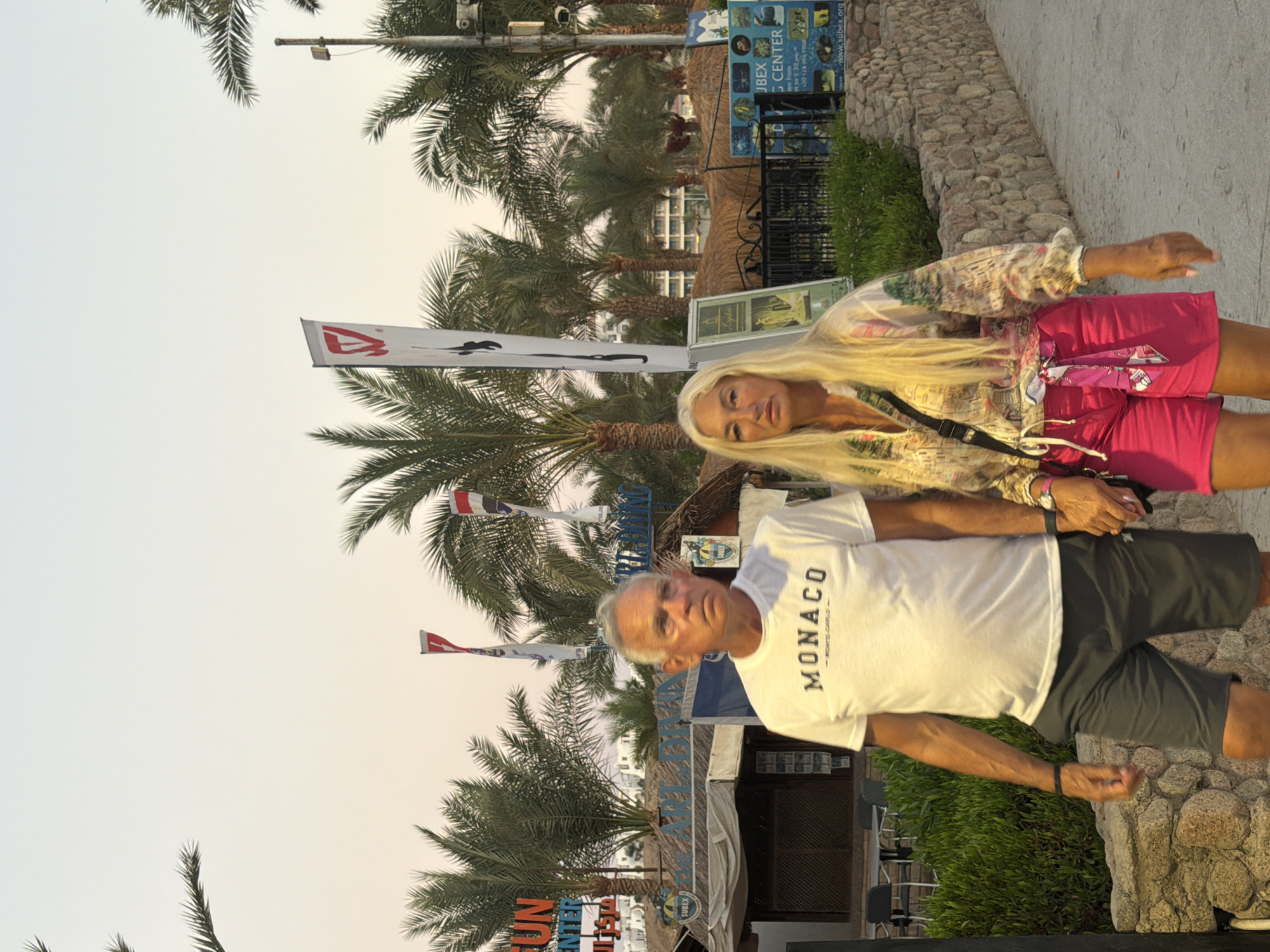 An older white man and an older white woman are walking outdoors in Sharm El Sheikh, Egypt. The man is wearing a white t-shirt with "MONACO" printed on it, dark gray shorts, and a black wristband. The woman is wearing a long-sleeved, multi-colored patterned shirt and bright pink shorts. She has a black crossbody bag and a pink scarf tied around her waist. They are holding hands and appear to be walking towards the viewer.
In the background, there are palm trees and a building complex with straw-covered roofs. Signs are visible, including one that says "FUN CENTER" and another that appears to be for a "SUBEX DIVING CENTER." The sky is a pale orange and yellow, indicating dusk or dawn. A stone wall lines the right side of the pathway.