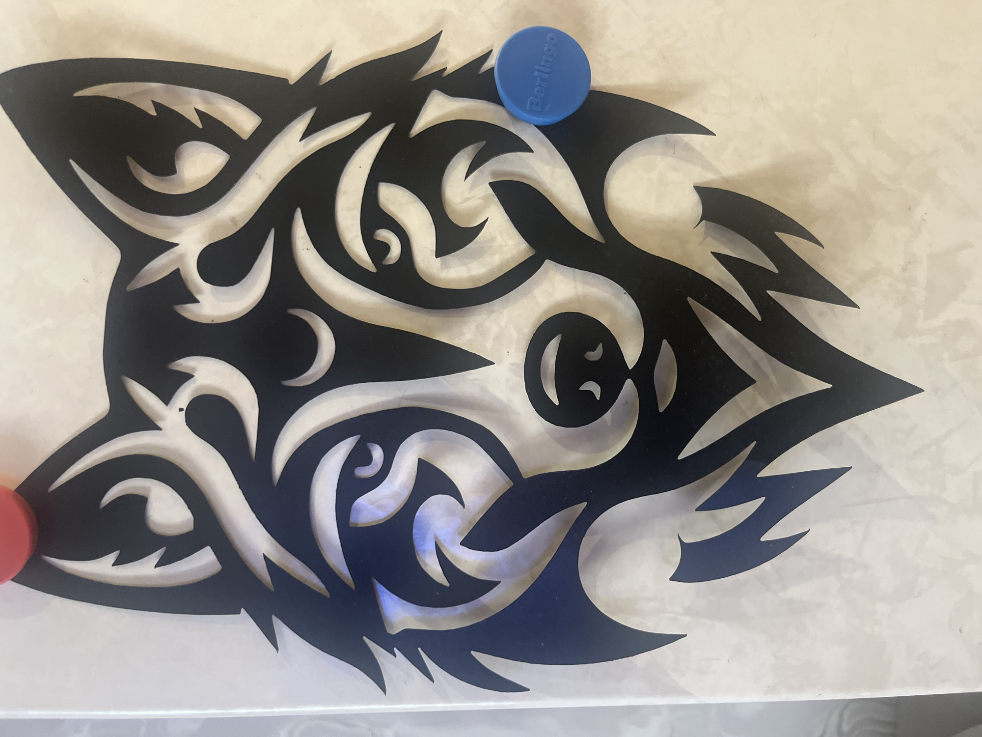 A black tribal wolf head design is affixed to a wall. The design is abstract, with sharp lines and curves that create the impression of a wolf's face. The background wall has a light, marble-like pattern. A red circular object is visible at the top of the image, partially obscured by the wolf's design. A small blue circular object with the word "Berlingo" printed on it is attached to the right side of the wolf's face. The scene appears to be indoors. There is no information to suggest the location is in the city of Tyumen, Russia.