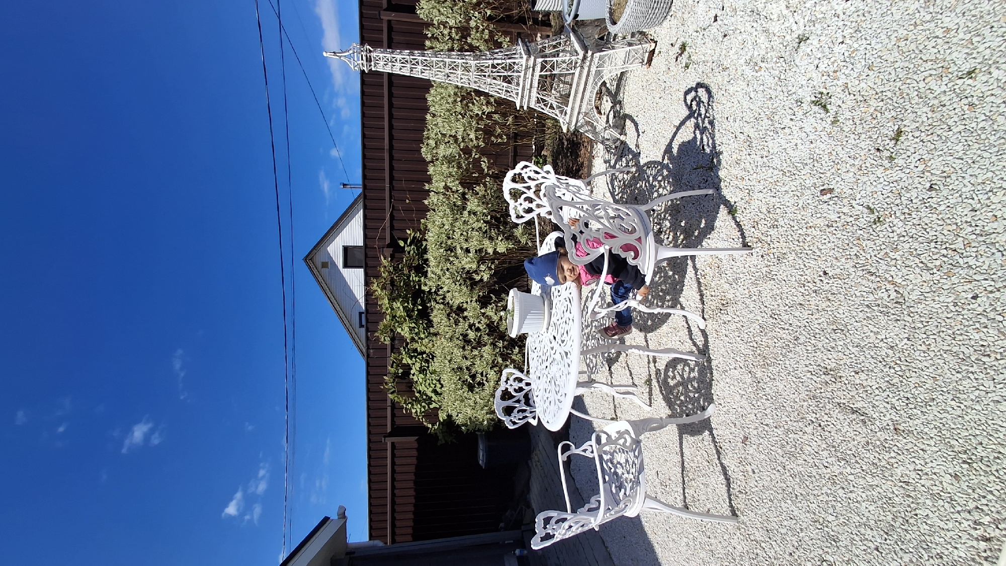 The image depicts an outdoor setting in what appears to be a backyard or patio area. A child is seated at a white metal table with ornate chairs. Behind the table and chairs, there's a brown fence or wall and a patch of greenery, possibly bushes or shrubs. A white Eiffel Tower replica stands behind the greenery. The ground is covered with small stones or gravel. The sky is blue with some clouds. The location might be Oliki, Russia, based on the question.