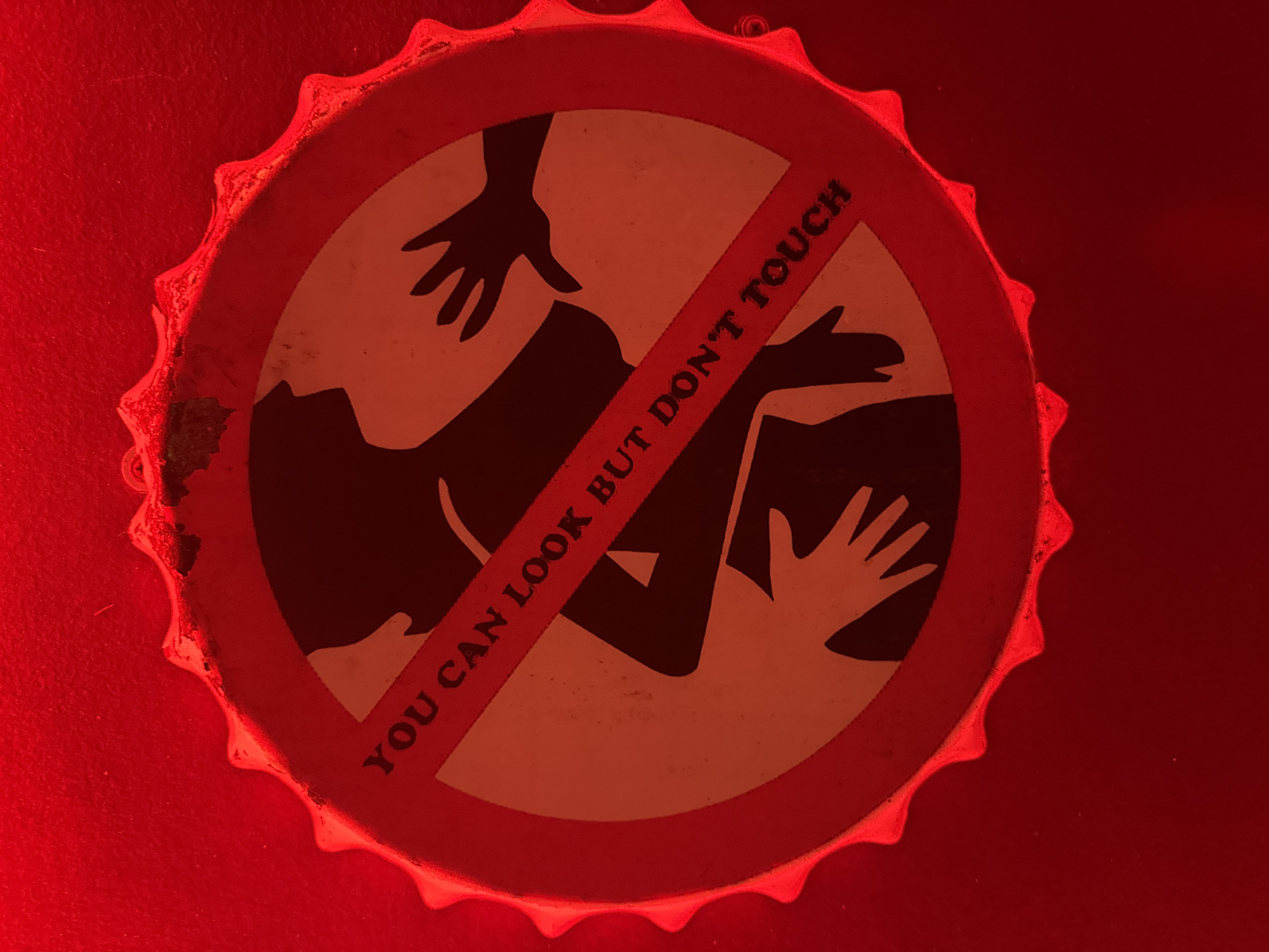 A red-toned close-up photograph captures a bottle cap featuring a controversial design. The cap is crimped at the edges and shows a circular logo with a crossed-out image of a suggestive scene involving two people and their hands. The text "YOU CAN LOOK BUT DON'T TOUCH" is written across the crossed-out area. The overall lighting gives a strong red hue to the object and the surface it rests on. The image has a strong red hue. It is not possible to verify the city of Đà Nẵng, Vietnam, as the location from the visual data.