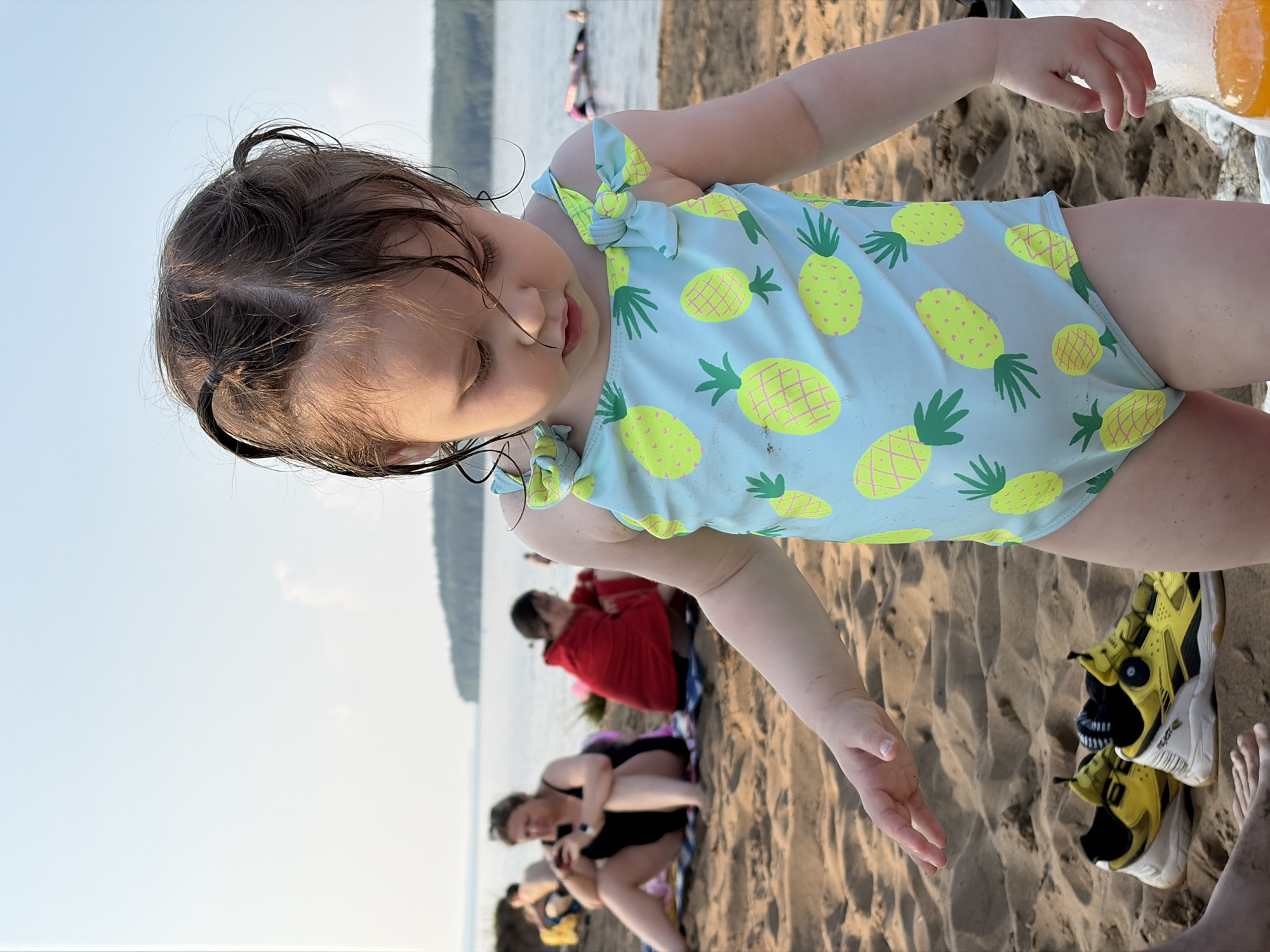 This is a medium shot of a baby girl wearing a light blue swimsuit with a pineapple pattern. She has wet, dark hair and is looking down with her eyes closed. The baby is on a sandy beach, with other people in the background. To the right of the baby are a pair of bright yellow and black sneakers.
In the background, several people are visible sitting on blankets on the sand, with a body of water and a distant shoreline in the far background. One person is wrapped in a red towel, and another is in a black swimsuit. The sky is clear and bright, suggesting it is daytime. The scene appears to be a casual outing at a beach or lake. The overall mood seems relaxed and summery. There is no visible text in the image.