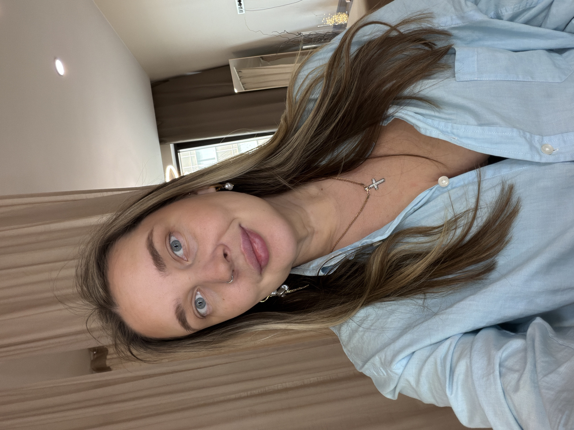 A woman with fair skin and light blue eyes is posing for a photo. She has light brown hair with blonde highlights. She is wearing a light blue button-down shirt. A gold necklace with a cross pendant is visible on her neck. She is wearing earrings and a nose ring.
There is no information to suggest that the photo was taken in Moscow, Russia.