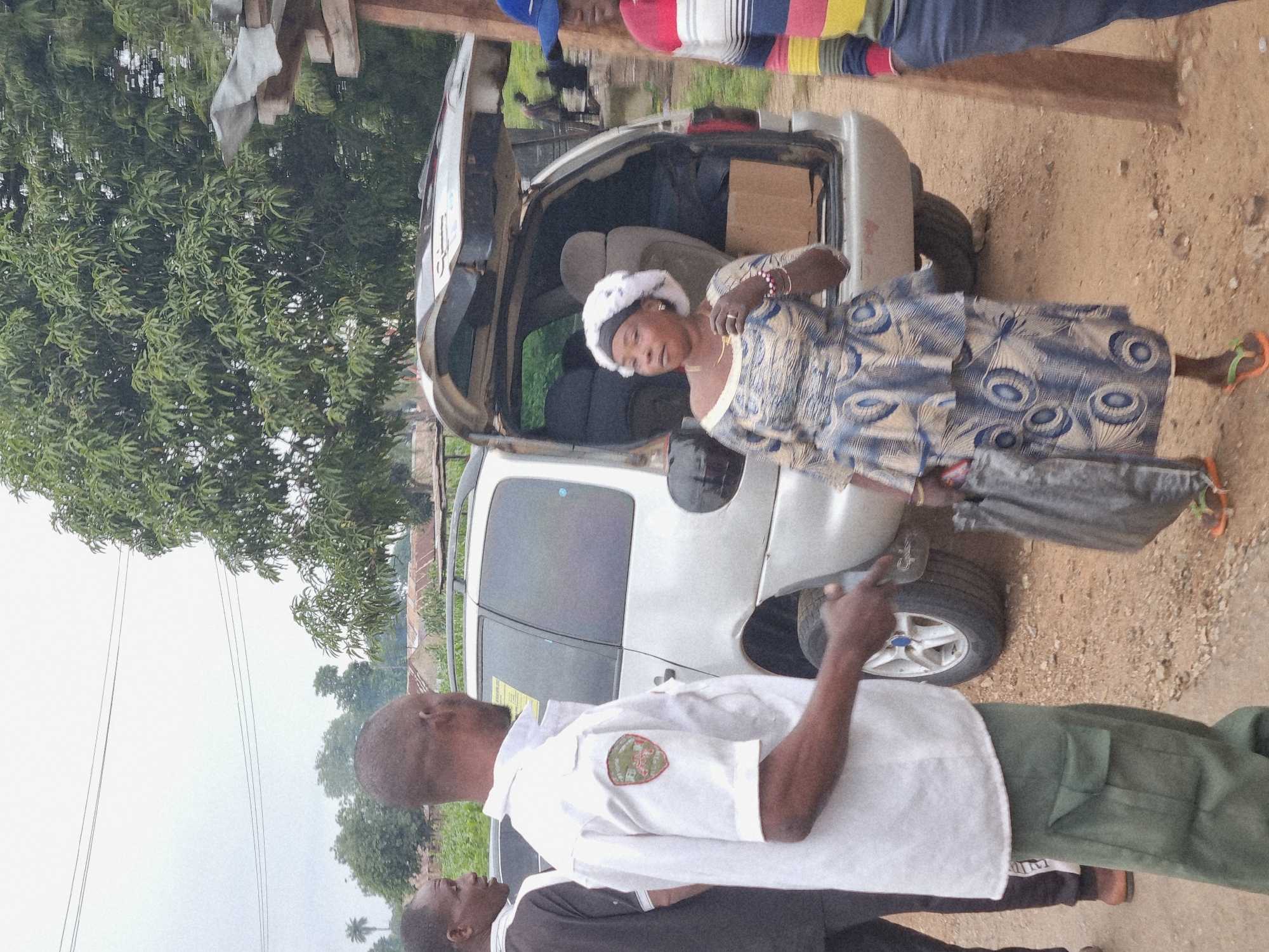 In the city of Marke, Nigeria, a group of people are gathered around a silver minivan with its rear hatch open. A man in a white short-sleeved uniform shirt with a patch on the left breast and green cargo pants stands in the foreground, facing away from the camera and gesturing with his left hand. To his left, another man in a black shirt and dark pants is partially visible. A woman in a blue and white patterned dress and a white head covering is standing to the right of the minivan, holding a dark bag. Her right hand is near her face, and she is wearing a necklace and bracelets. Behind her, a person in a striped shirt and blue pants is visible. The ground is covered in gravel and dirt, and vegetation and buildings are visible in the background under an overcast sky.
