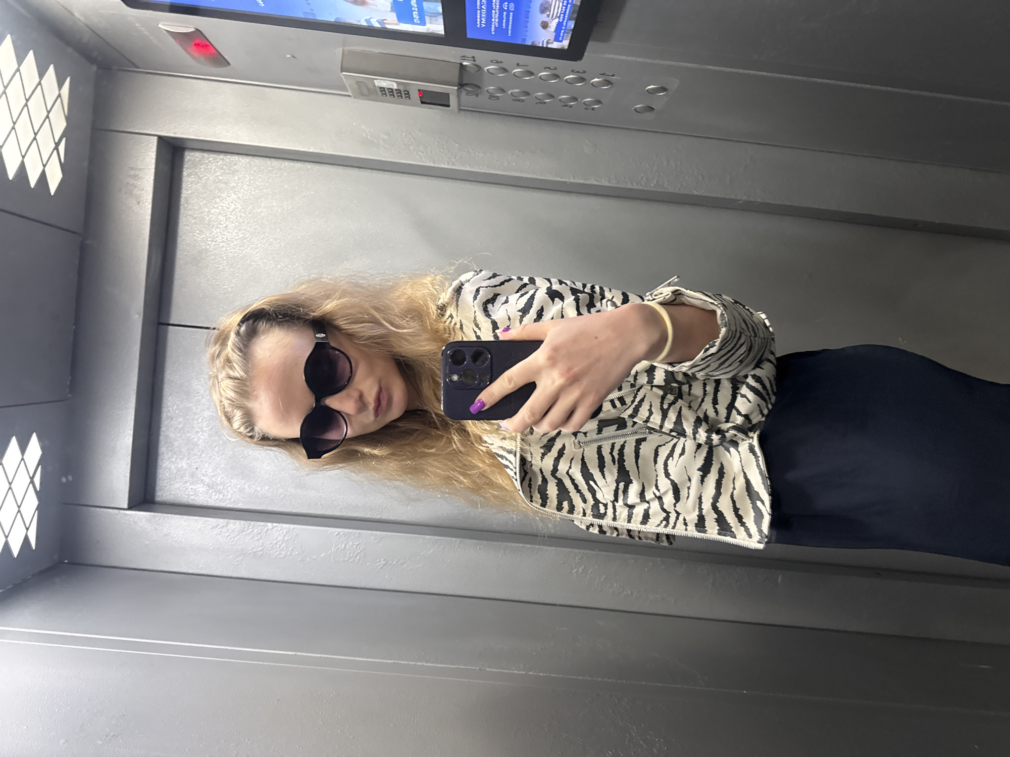 The photograph captures an individual with long blonde hair, wearing large sunglasses and a zebra-striped jacket, taking a selfie with a black smartphone. The person is also wearing a gold-colored bracelet on their left wrist and has purple nail polish on the hand holding the phone. The setting appears to be inside an elevator, judging by the control panel with numerous buttons in the upper portion of the image, as well as the elevator lighting and patterned ventilation grilles on the left side. The walls are a plain gray color.