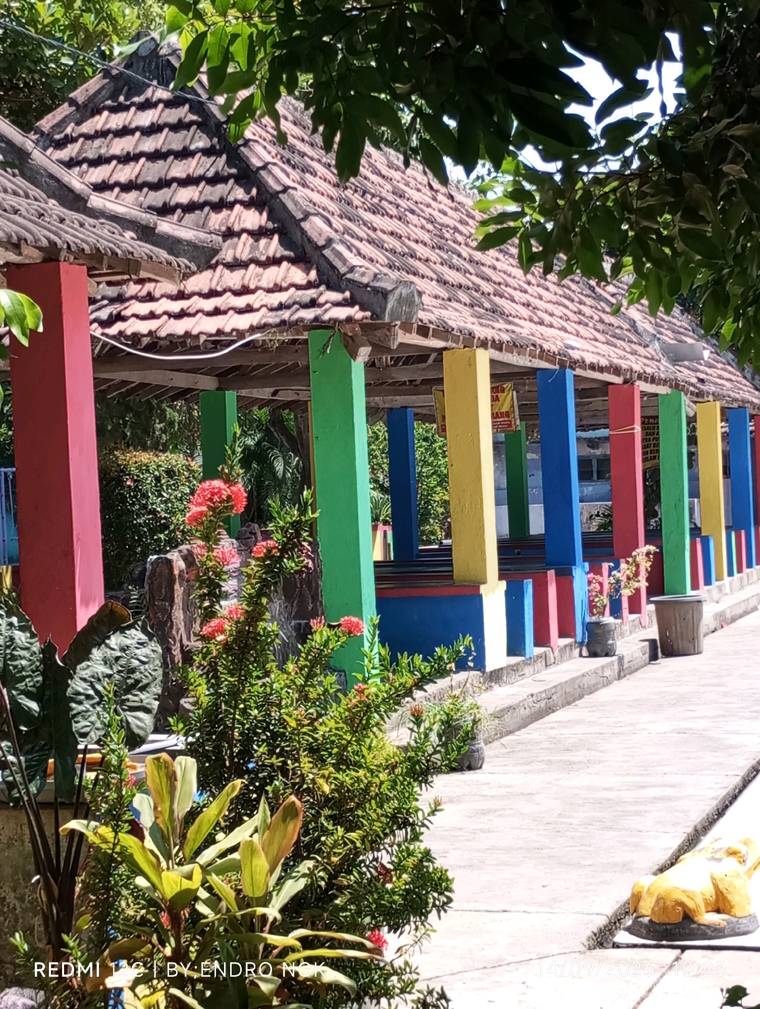The image depicts a covered outdoor seating area with a tiled roof supported by brightly colored pillars in red, green, yellow, and blue. The seating area appears to be an open pavilion, with a concrete walkway in front of it. Lush green vegetation, including flowering plants, surrounds the structure. In the foreground, there are large tropical-looking leaves and smaller plants. To the right of the walkway, a yellow decorative statue resembling a lounging animal is visible. The scene is illuminated by natural sunlight, casting shadows. The overall impression is one of a tropical, possibly recreational, outdoor space. The text overlay indicates the image was taken in Sombron, Indonesia, and captured with a REDMI 12c phone on 14/09/2026 at 10:23.