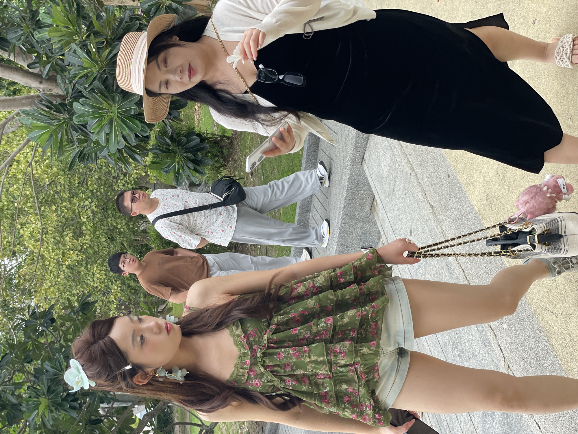 In a city environment in Na Chom Thian, Thailand, three individuals are depicted walking on a paved walkway with lush green trees and foliage in the background. The foreground is dominated by two women. The woman on the left is younger, wearing a floral-print green camisole with ruffled tiers and light-wash denim shorts. She has long, dark wavy hair adorned with a light blue flower and matching earrings, and she is carrying a black handbag with a chain strap and a pink fluffy charm. The woman on the right is older, wearing a black dress, a white cardigan, and a straw hat. She is holding a smartphone in her right hand and a white flower in her left. Two men are visible in the middle ground, walking behind the women. One man in a brown t-shirt and light gray pants, the other in a white printed shirt, dark pants, and a black crossbody bag. The ground is a textured concrete surface with stone steps in the immediate foreground.