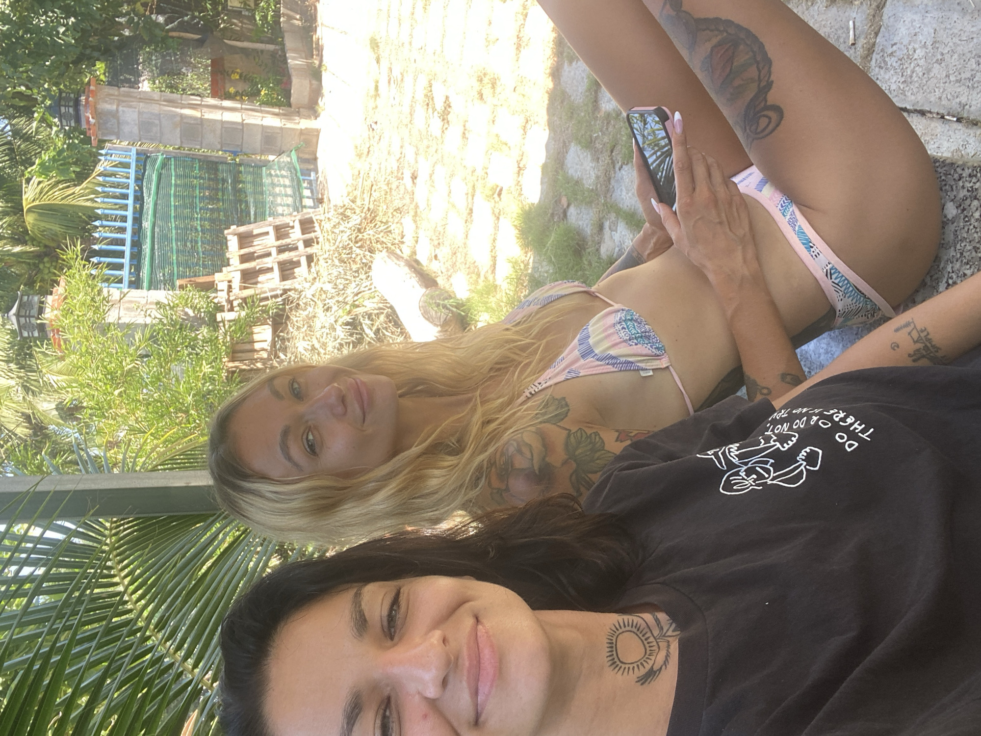 Two women are sitting outdoors in Mũi Né, Vietnam. The woman on the left has dark hair and is wearing a black t-shirt with a graphic print. She has a tattoo on her neck that resembles a sun. The woman on the right has blonde hair and is wearing a bikini. She has tattoos on her arms and legs. She is holding a smartphone in her hands. The background shows lush greenery, palm trees, a blue fence, and a stone wall. The ground is paved with stones.