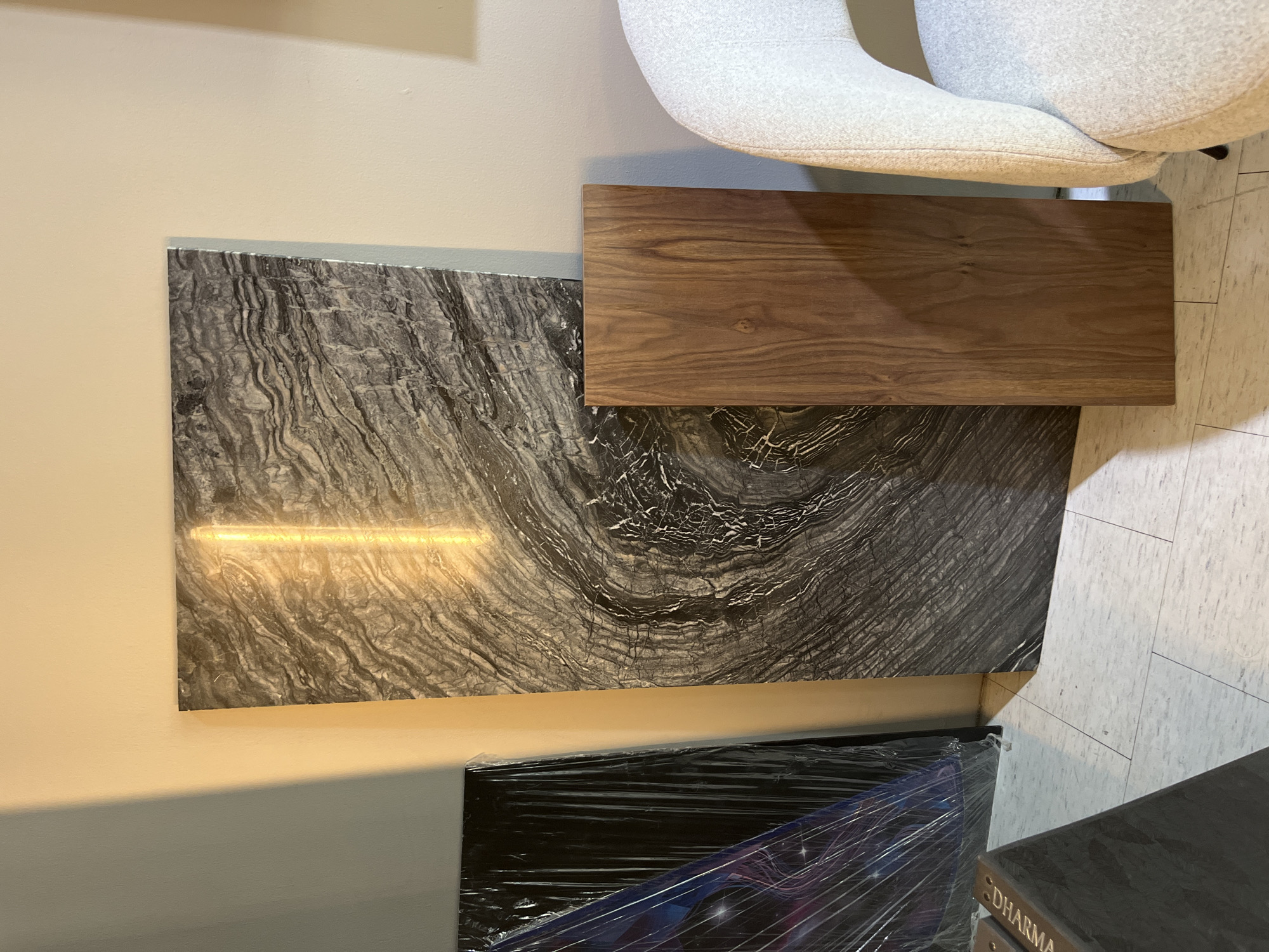 This image depicts an interior scene, likely a showroom or storage area, featuring several large rectangular slabs leaning against a wall. The primary subject is a dark, veined stone slab with dramatic swirling patterns, illuminated by a vertical strip of warm yellow light that runs through its center. To the left of this stone slab is another slab wrapped in clear plastic, revealing a dark background with what appears to be a starry or cosmic design. To the right of the main stone slab is a solid, medium-brown wooden slab with visible grain patterns. In the far right of the frame, a portion of a white, textured armchair is visible. The floor is tiled with light-colored, rectangular tiles. The wall behind the slabs is a neutral, light color, with a distinct shadow cast from an unseen light source on the left side. The overall impression is one of organized display or storage of large material samples or finished products. The location in San Bernardino Tlaxcalancingo, Mexico, is not discernible from the visual elements of the image itself.