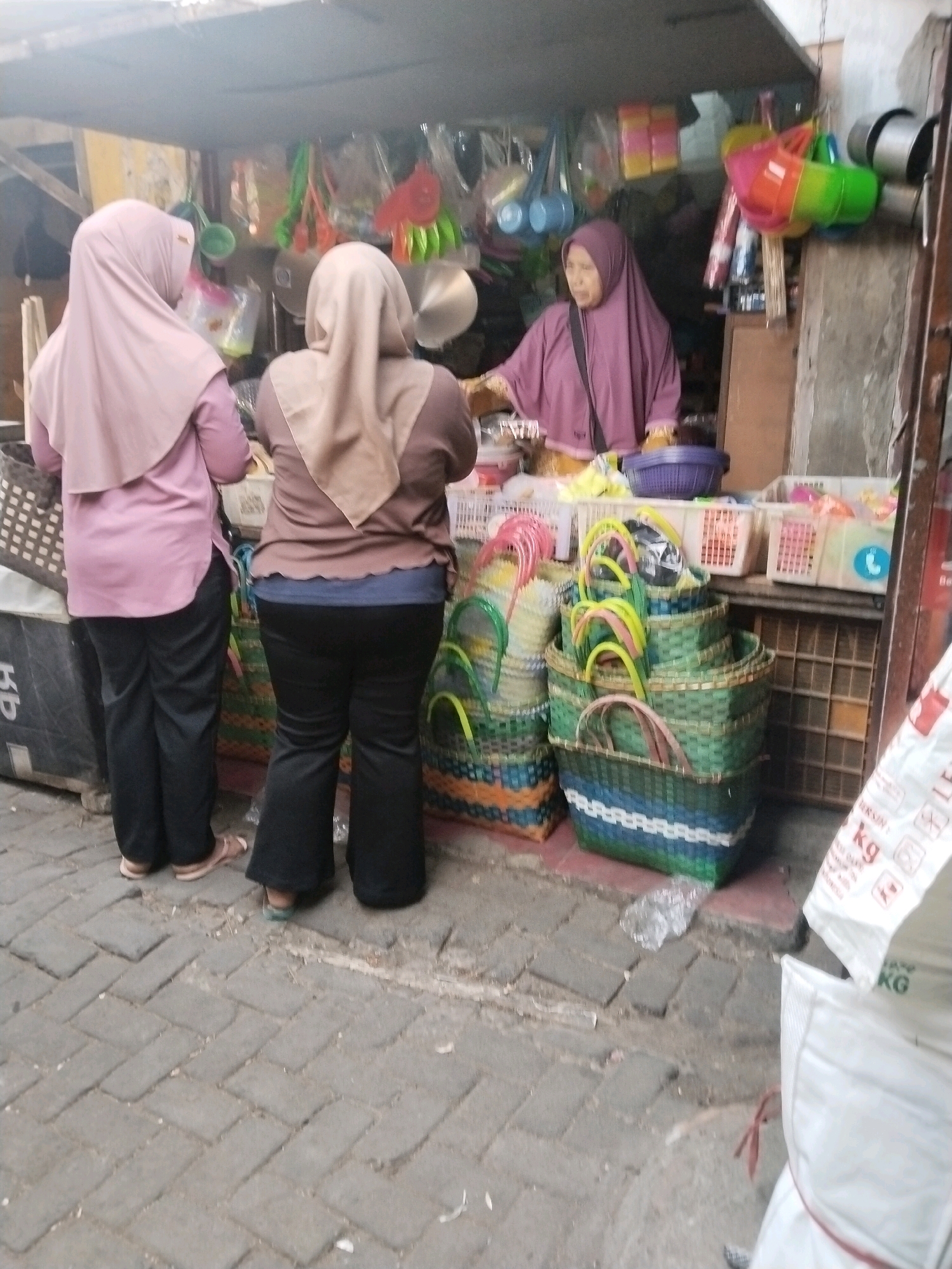 In Pandanarum, Indonesia, a street market scene unfolds. Three individuals are present at a vendor stall, which appears to be set up under a simple awning. Two women are positioned facing away from the camera, interacting with a vendor who is an older woman wearing a purple hijab. The stall is filled with various items, predominantly woven baskets in different sizes and colors, stacked in multiple rows. Other items visible include plastic containers, what seem to be kitchen utensils like ladles, and various colorful plastic goods hanging from the awning. The ground is paved with interlocking bricks. The overall impression is one of a bustling local marketplace.